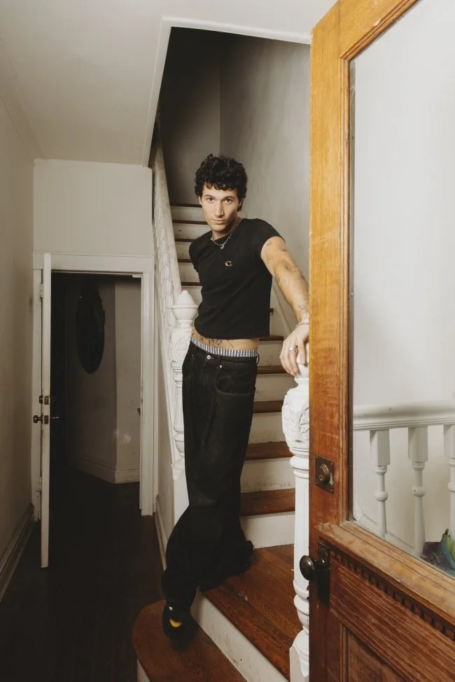 Young man with curly hair wearing a black t-shirt and black jeans, leaning on a staircase banister in a house interior.