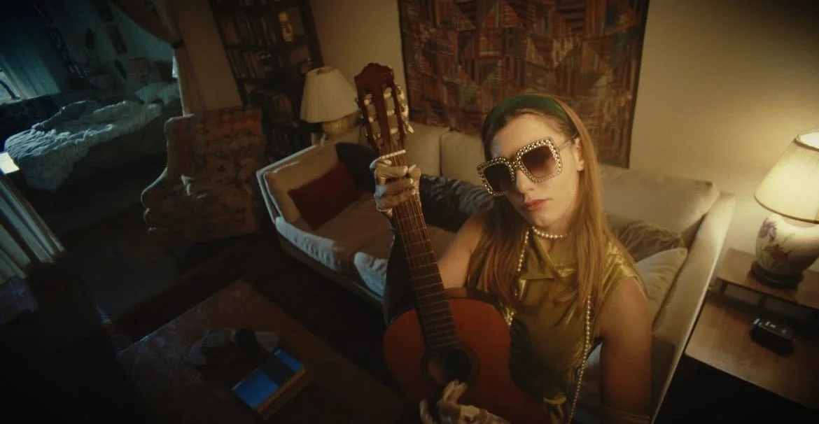 A woman with long red hair wearing large glasses, a pearl necklace, and a gold top, holding an acoustic guitar in a cozy living room.
