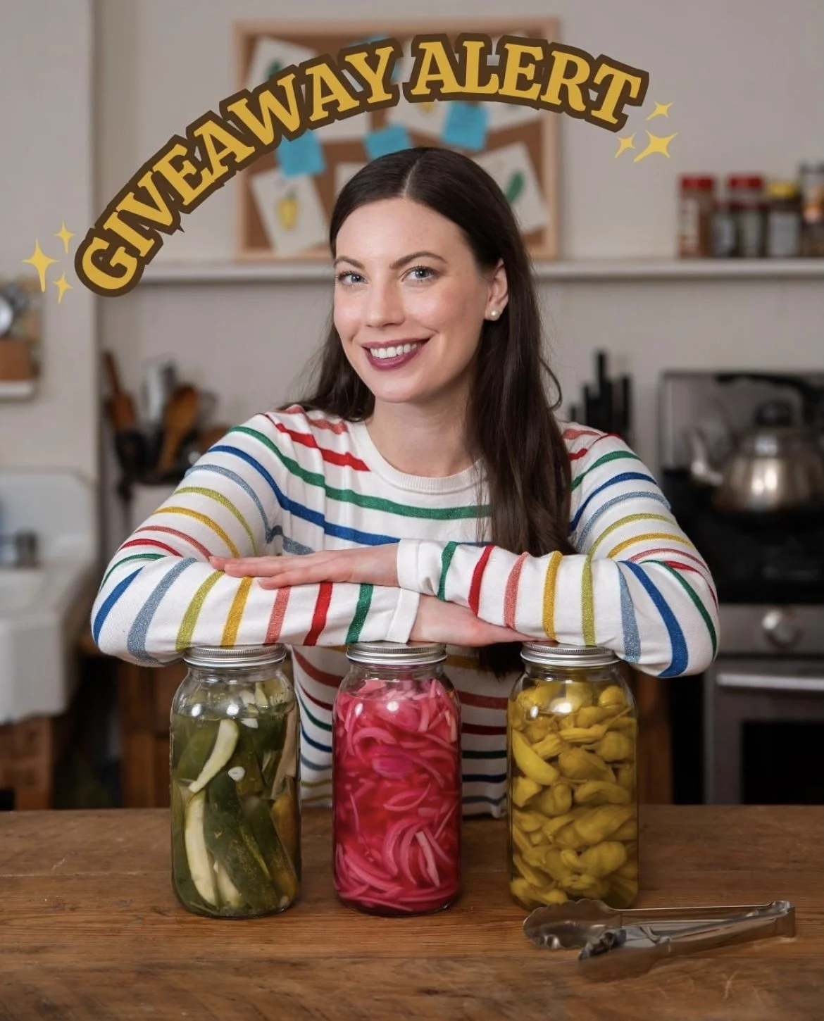 Woman in a rainbow-striped sweater smiling behind three jars of pickled vegetables on a wooden table in a kitchen.