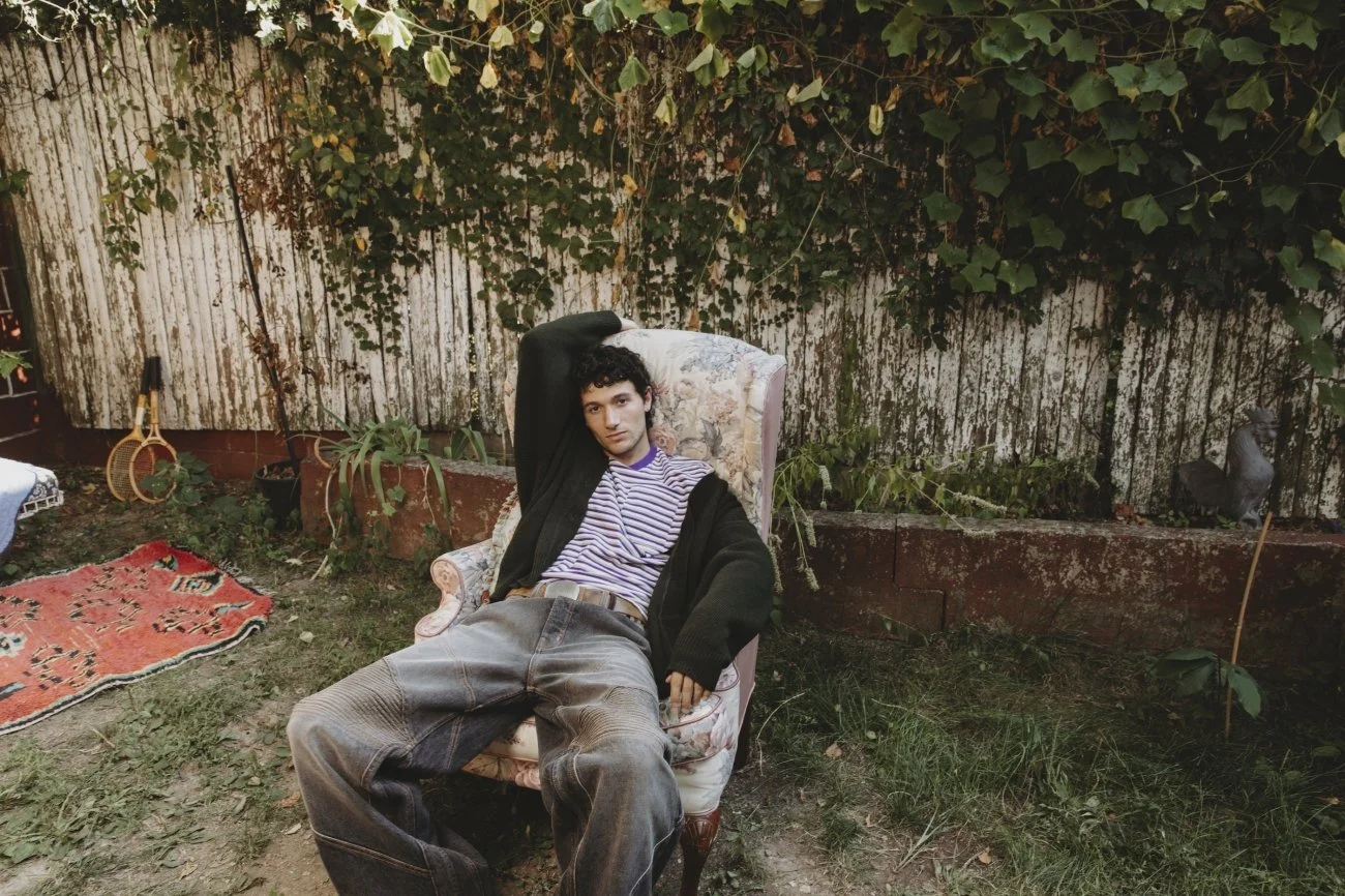 A young man with curly hair wearing a striped shirt, black cardigan, and baggy jeans, relaxing on an old floral armchair outdoors. The setting is a backyard with overgrown grass, plants, and a weathered wooden fence covered with ivy. There is a red p