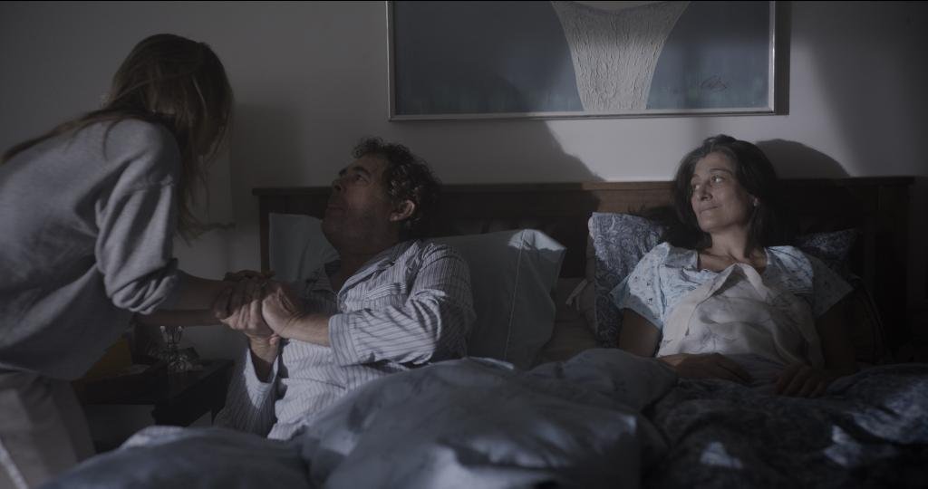 A woman in a hospital bed holding a man's hand, another woman sitting on the bed looking at them, in a dimly lit room.