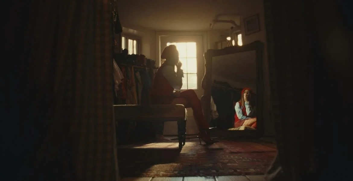 A woman sitting on a bench inside a dimly lit room, looking at her reflection in a large standing mirror, with sunlight coming through a window in the background.