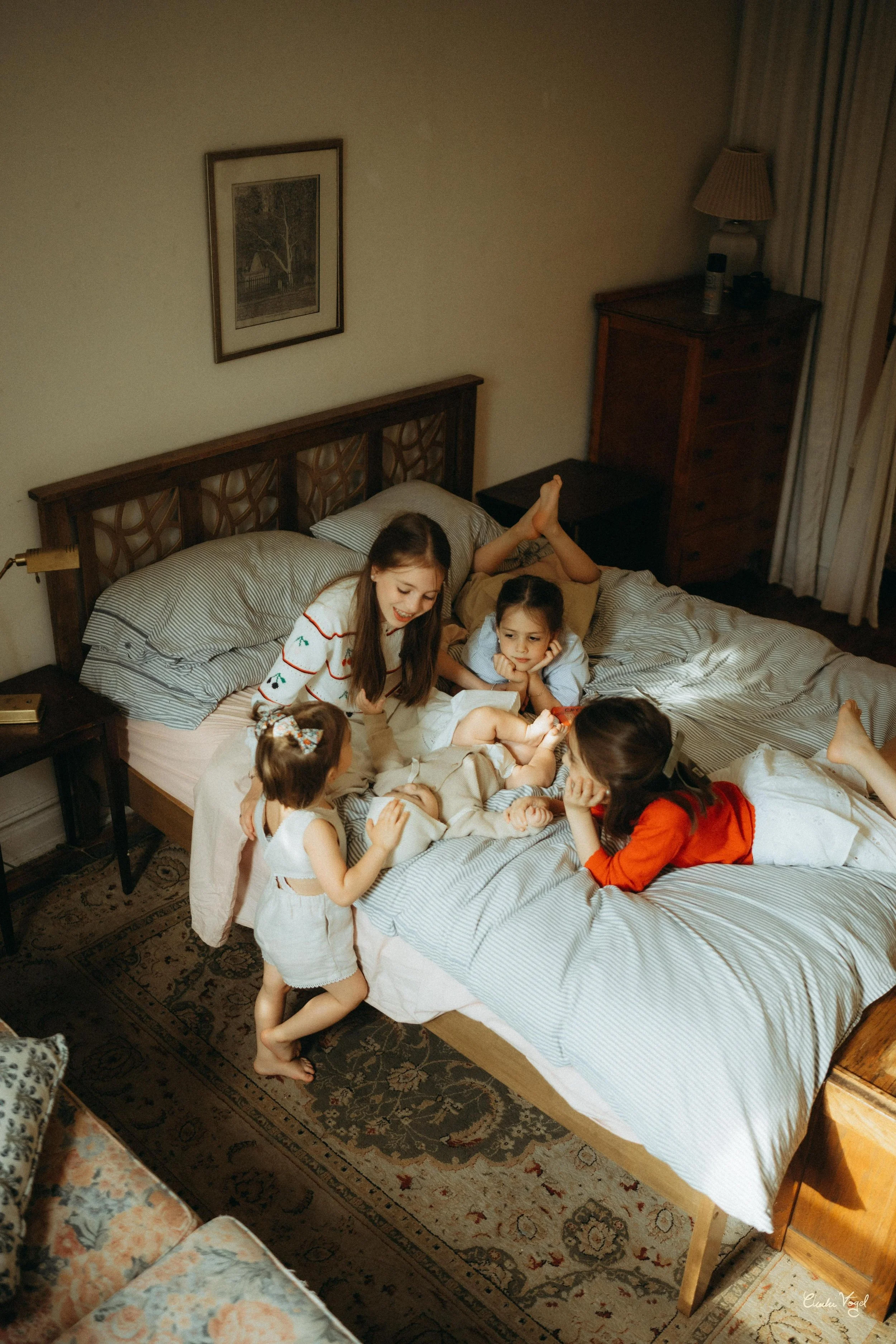 A group of children and a young woman are sitting on a bed, looking at a baby who is lying down and holding a red toy. The children appear engaged and happy, in a warmly lit bedroom with a wooden bed frame, framed pictures on the wall, and curtains.