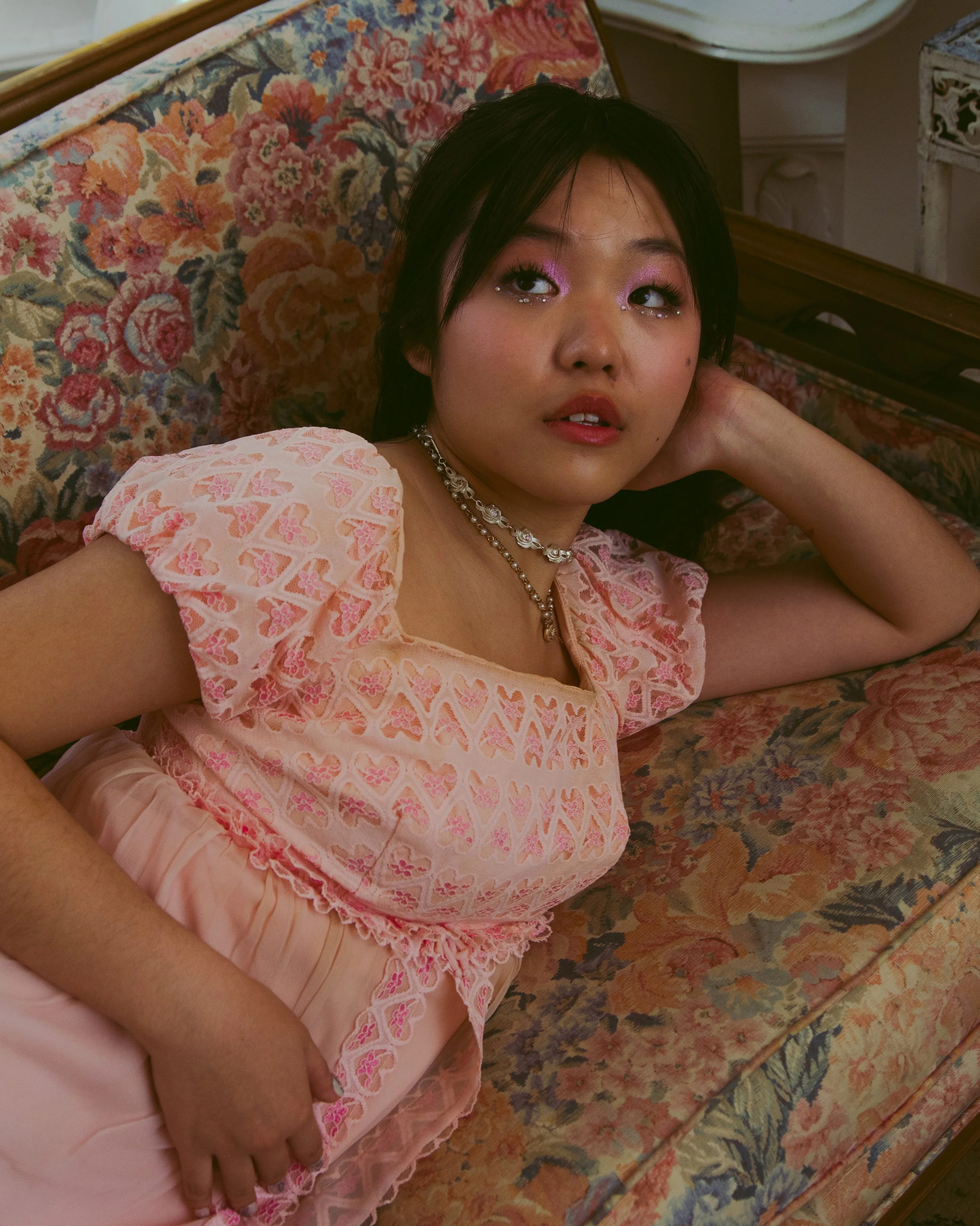 Young woman lying on a floral-patterned vintage sofa, wearing a pink dress with lace and heart motifs, accessorized with layered necklaces, and pink eyeshadow, looking away with a contemplative expression.