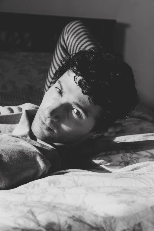 Young man with curly hair lying on a bed, in black and white, with one arm behind his head and reading light casting shadows on his face.