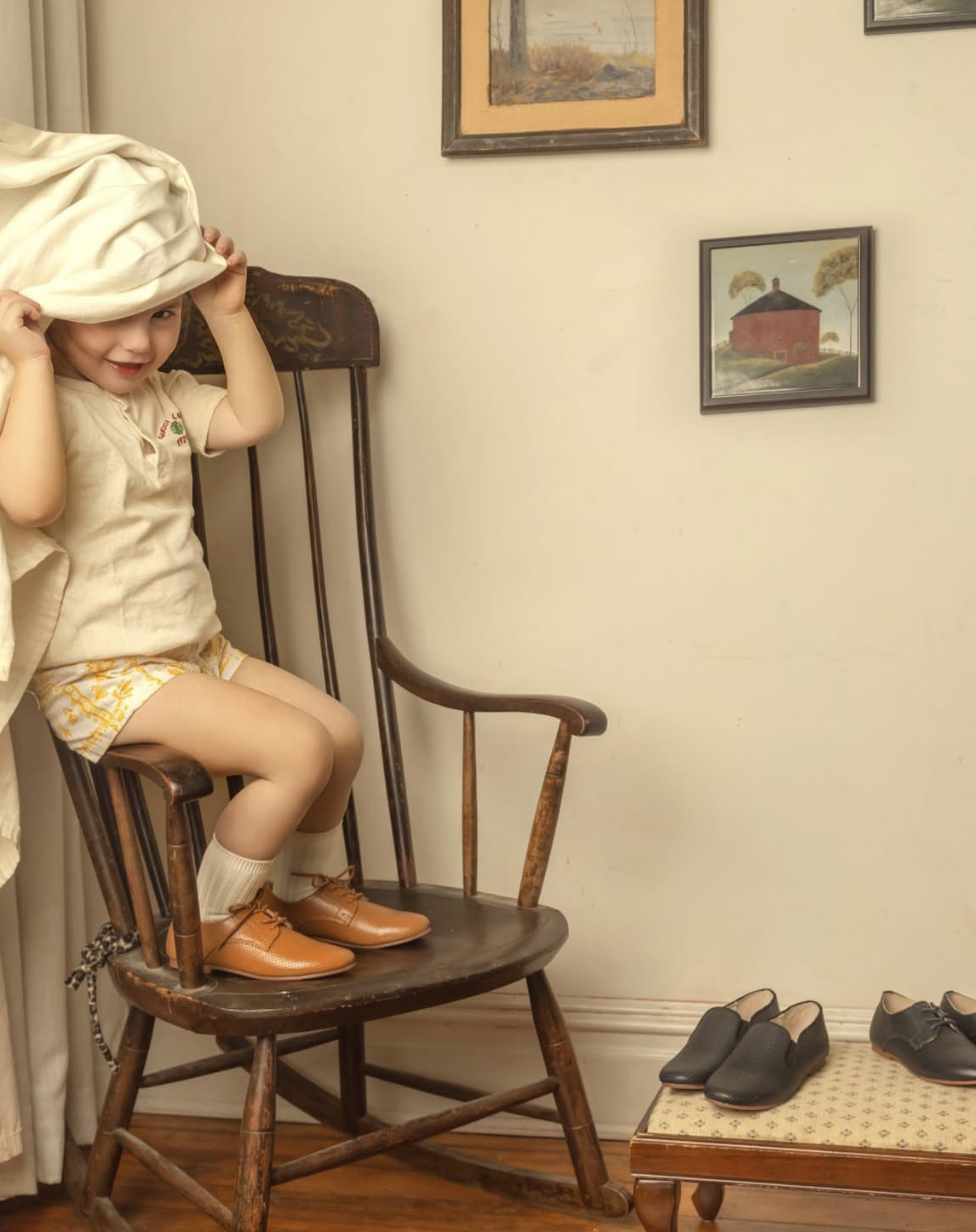 A young girl sitting on a wooden rocking chair, putting on or taking off a large cream-colored hat. She is wearing a beige shirt, yellow shorts, cream socks, and brown shoes. The room has framed landscape artwork on the wall and a small cushioned sea