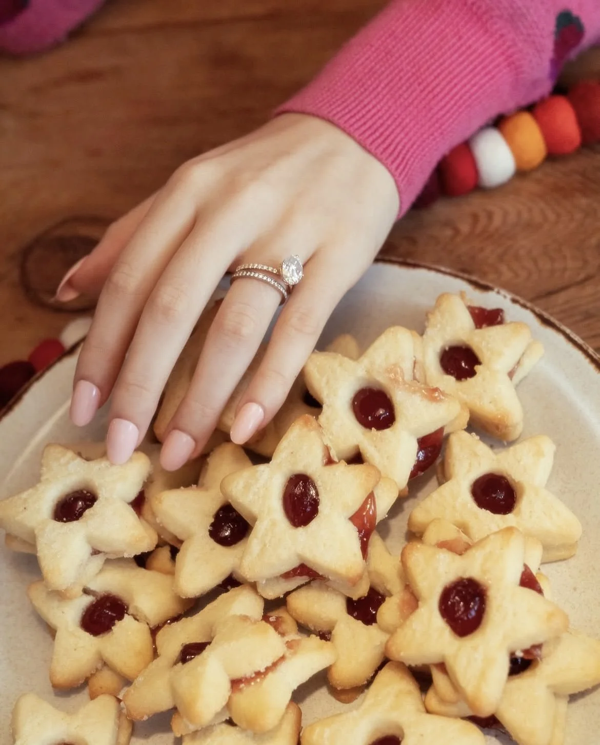 A person's hand with pink nail polish, wearing a silver ring with a large gemstone, resting on a plate of star-shaped thumbprint cookies with red jelly centers, on a wooden table.