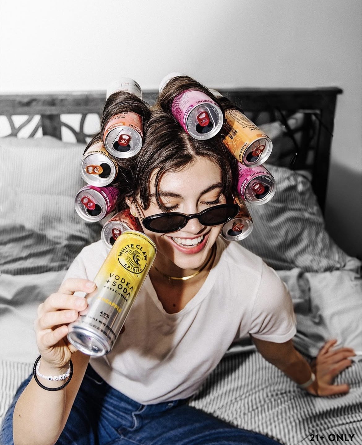 Woman with hair styled using soda cans, holding a vodka and soda can, smiling and wearing sunglasses.