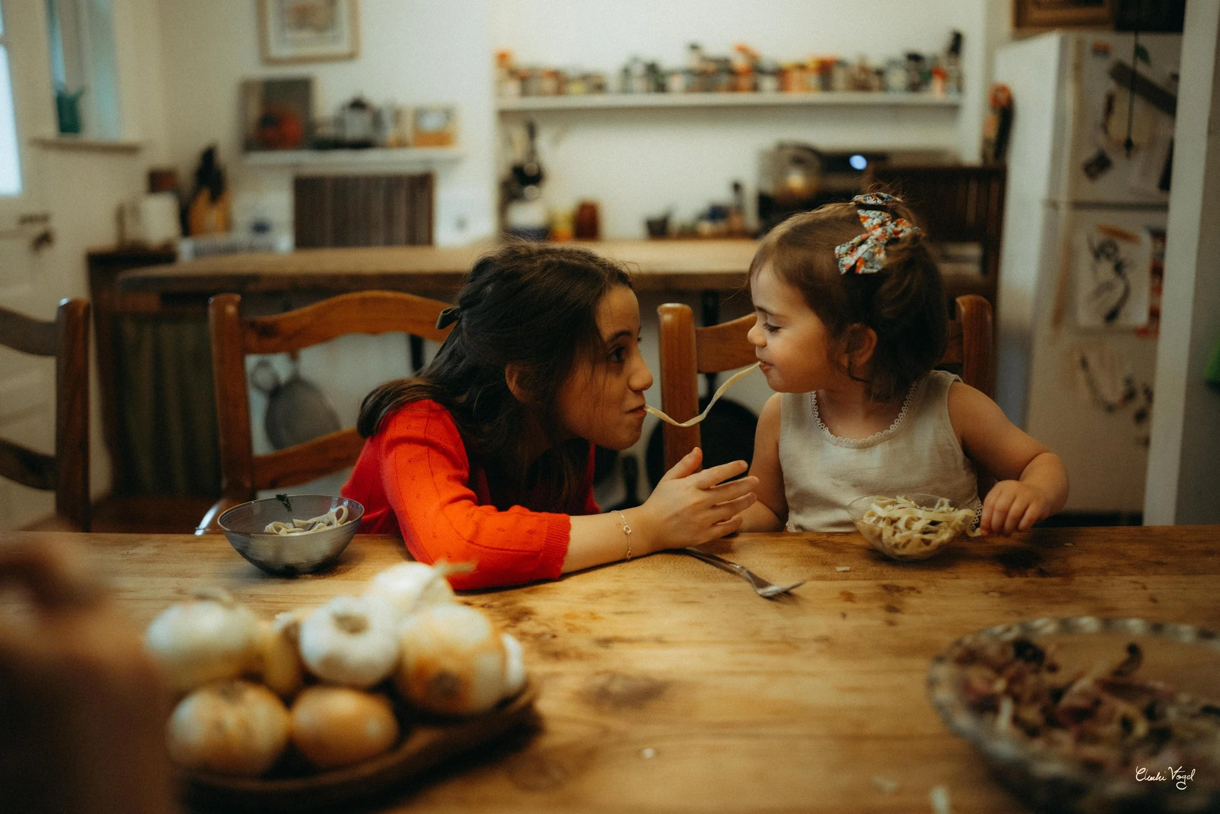 A woman and a young girl sharing a spoon of pasta at a wooden dining table. The woman is wearing a red top and the girl has a white sleeveless shirt with a blue bow in her hair. There are bowls of pasta and garlic bread on the table.