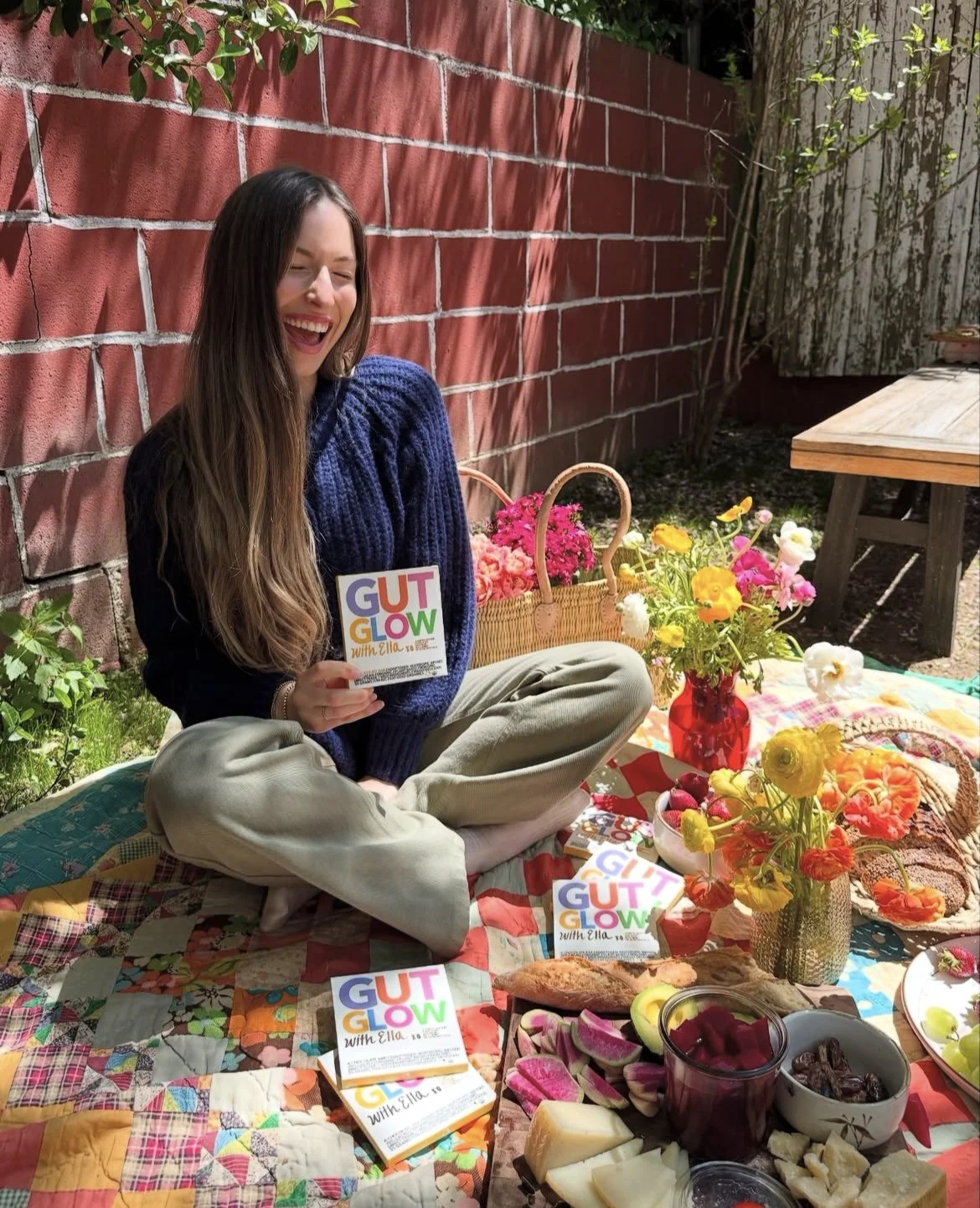 Woman sitting on a patchwork quilt in a backyard, laughing and holding a book titled 'Gut Glow with Ella'. There are flowers, a picnic basket, and food items on the quilt, with a red brick wall and greenery in the background.