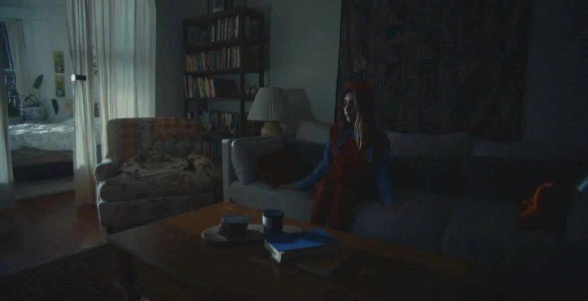 A woman with red hair sitting on a dark gray couch in a dimly lit living room, with bookshelves, a lamp, and a tapestry on the wall behind her.