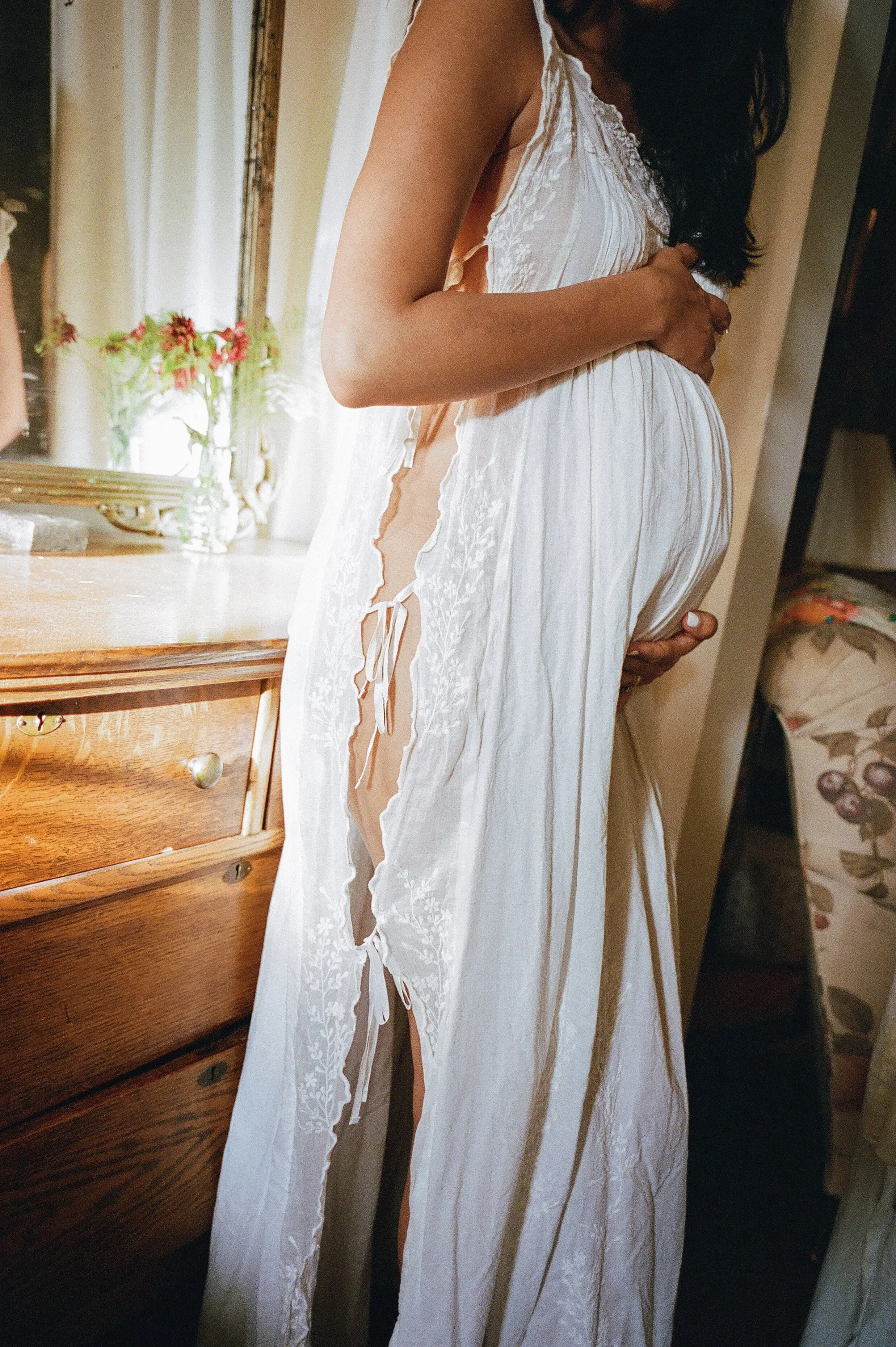 A woman in a white, lace-trimmed dress with side cutouts, standing indoors near a wooden dresser and a mirror, holding her pregnant belly.