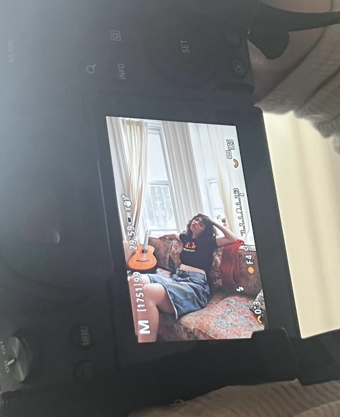 A camera's live view screen shows a young woman sitting on a vintage floral sofa with a guitar on her lap, wearing an oversized black T-shirt and denim skirt, in a room with a window and curtains in the background.