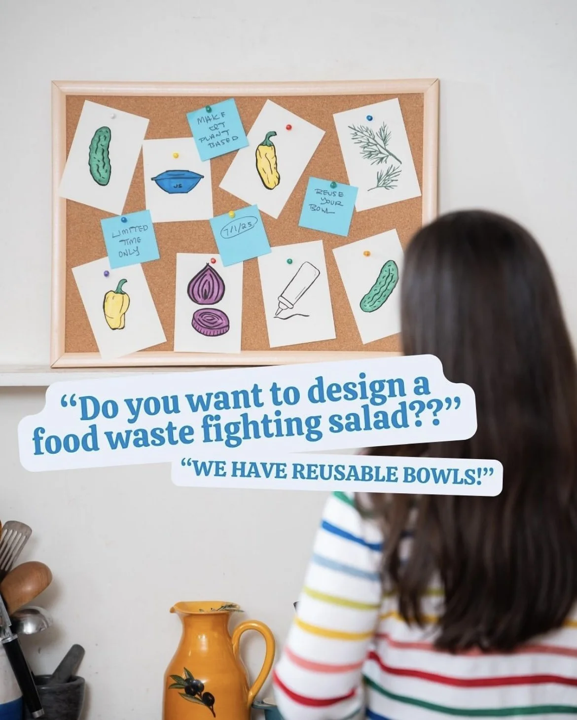 A person with long dark hair and a striped multicolored shirt looks at a debate about food waste and reusable bowls displayed on a bulletin board. The board has various drawings of vegetables and notes, and there is a quote about designing a food was
