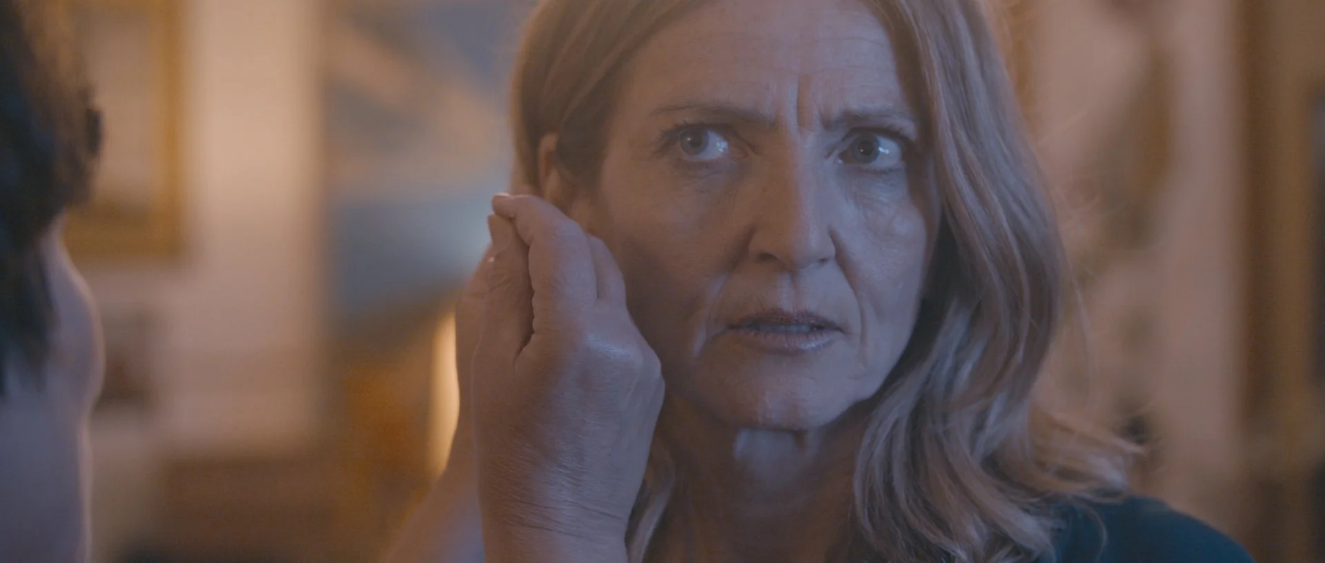A woman with long hair looks to the side with a serious expression as an older woman's hand touches her cheek.
