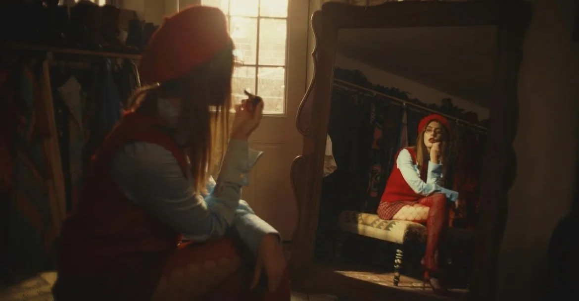 A woman with red hair and glasses is sitting in front of a large mirror with a carved wooden frame. She is wearing a red beret, a red vest, fishnet tights, and high heels. The room is warmly lit, with a window behind her letting in natural light. The