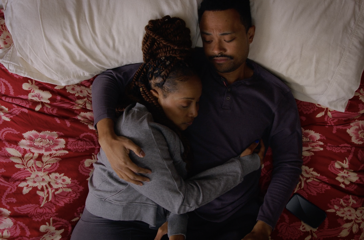A couple lying in bed, embracing and appearing to be sleeping or resting, with floral patterned bedding and pillows.