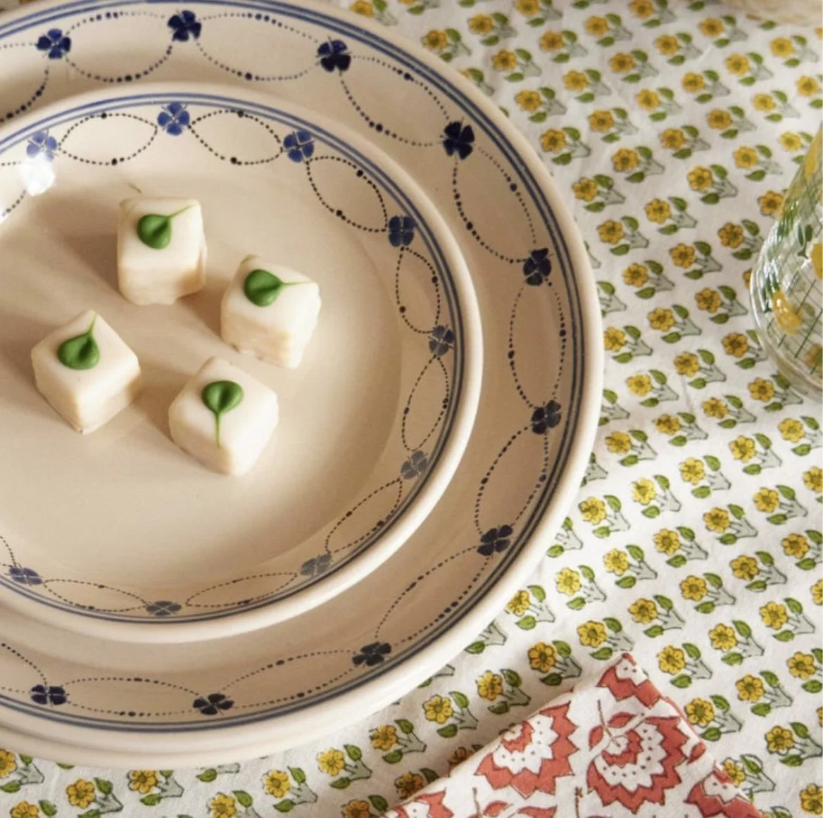 Four small white candies topped with green leaves on a patterned china plate, placed on a floral tablecloth with a glass vase to the side.