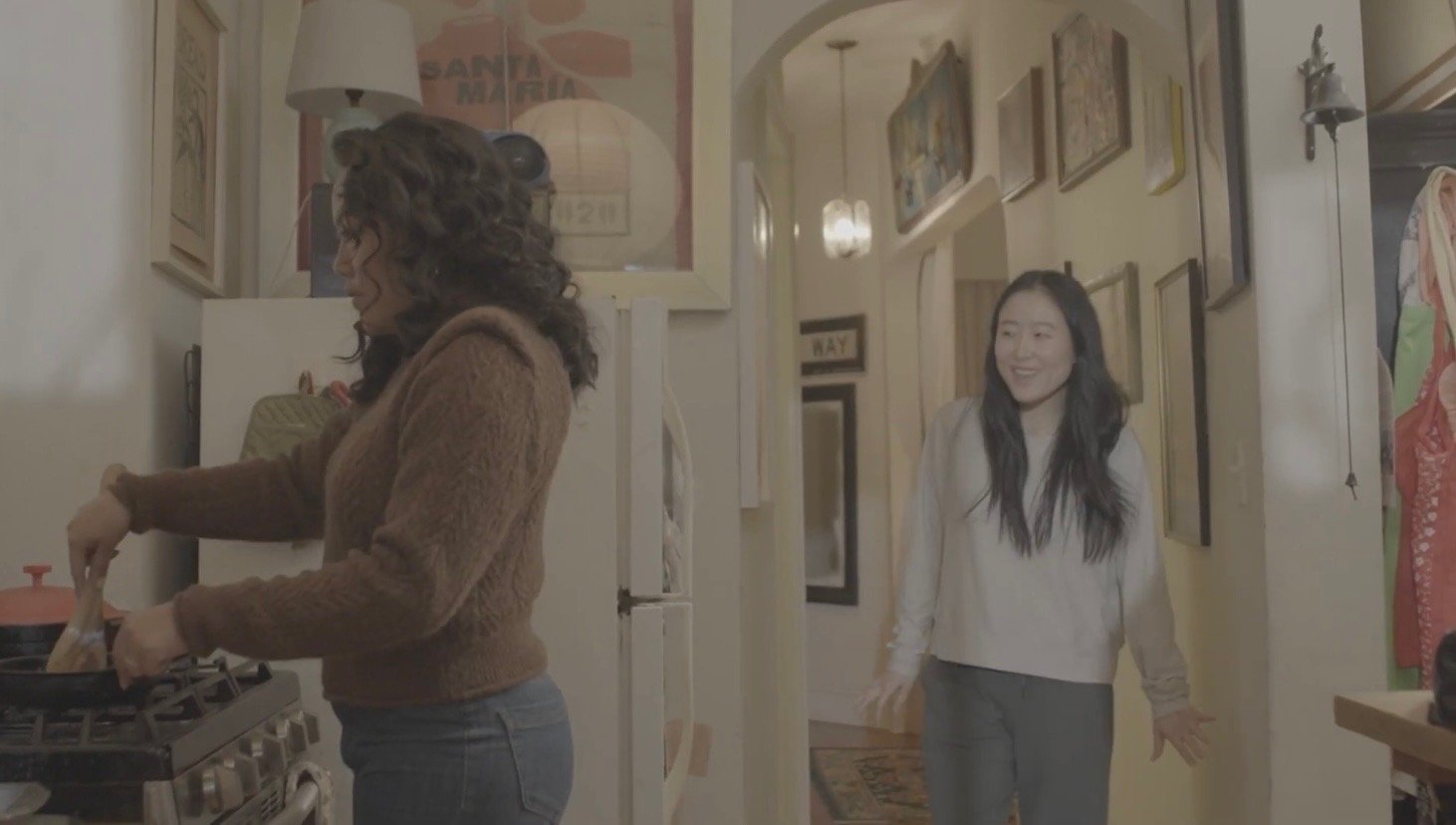 Two women in a cozy kitchen, one cooking at the stove and the other smiling in the doorway.