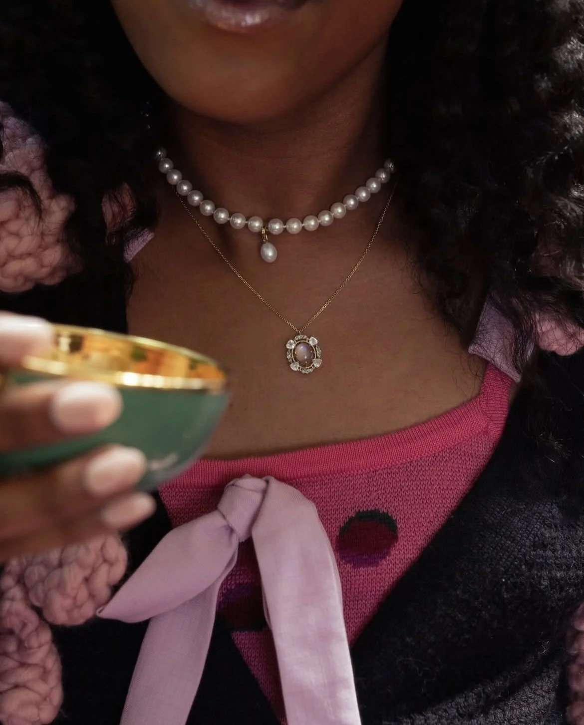 Close-up of a woman wearing pearl necklaces and a pink blouse with a bow, holding a small round container with a gold rim.