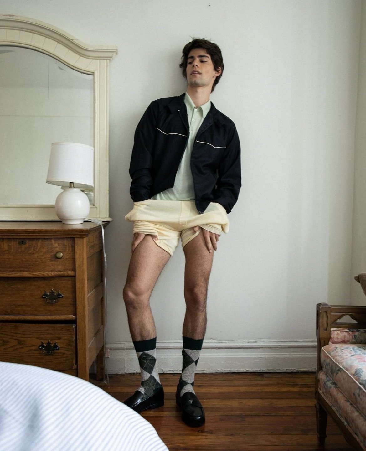 Young man standing in a bedroom, wearing a dark jacket, light shirt, yellow shorts around his ankles, argyle socks, and loafers.