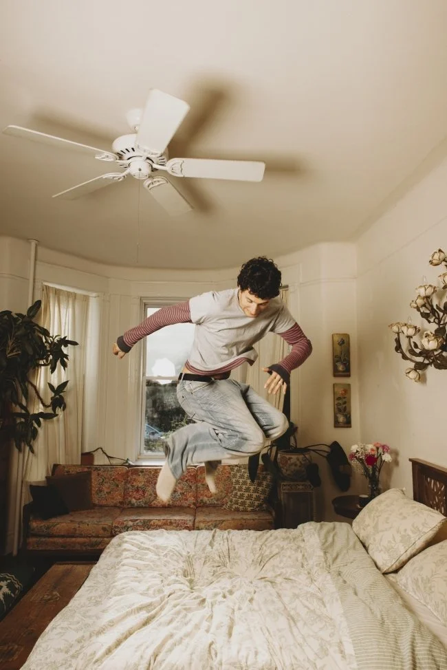 Person jumping on a bed in a bedroom with a ceiling fan, window, couch, plants, and floral wall decor.