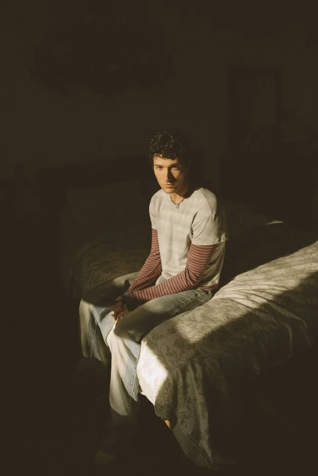 A young person with short, dark, curly hair sitting on the edge of a bed in a dimly lit room, wearing a white shirt with red and white striped long sleeves underneath, looking slightly towards the camera.