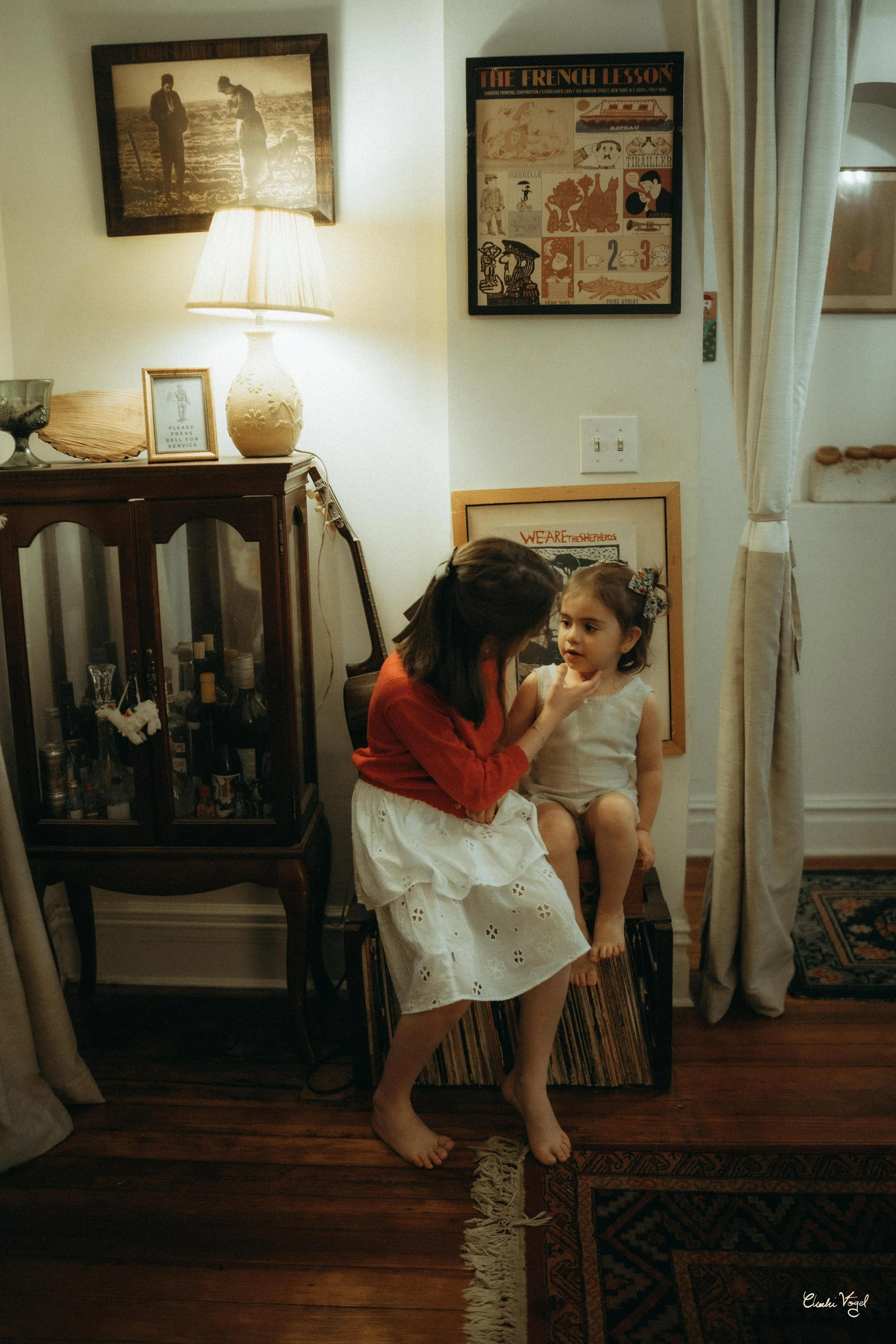 A woman and a young girl sitting on a cabinet, with the woman gently touching the girl's face in a cozy, vintage-style living room.
