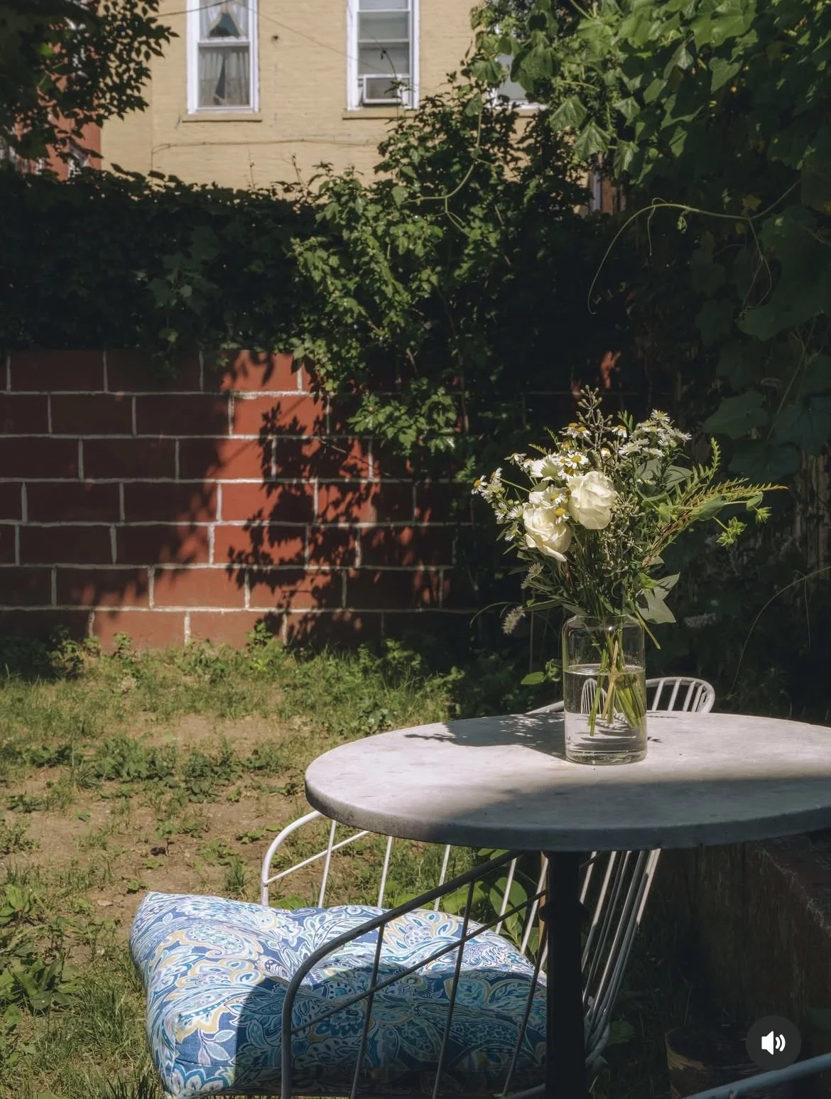 A vase of white flowers on a round outdoor table with a cushioned chair, in a garden with a brick wall and a yellow building in the background.