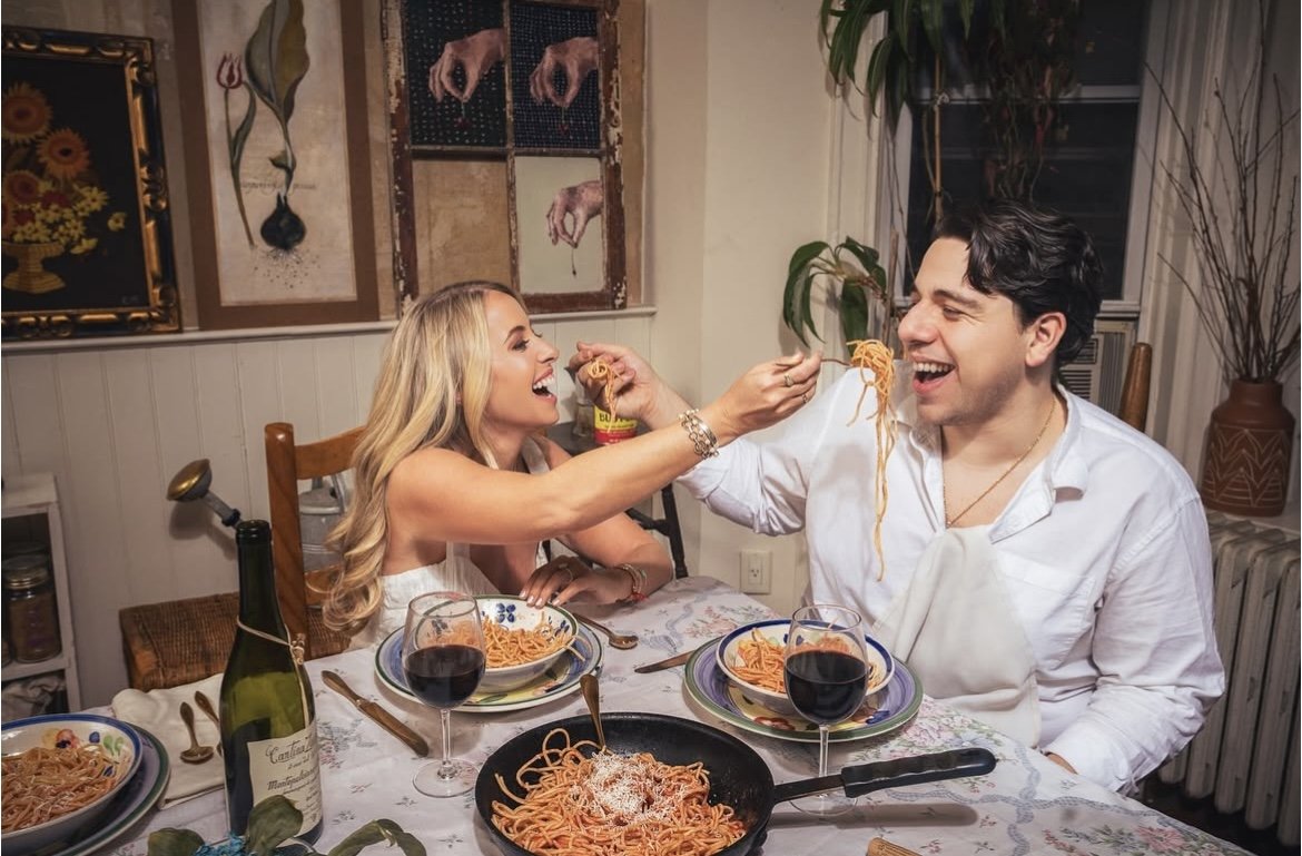 A couple sharing a fun moment at dinner, with spaghetti. The woman is feeding the man with spaghetti, both smiling and laughing, seated at a table with wine glasses, a bottle of wine, and plates of spaghetti.