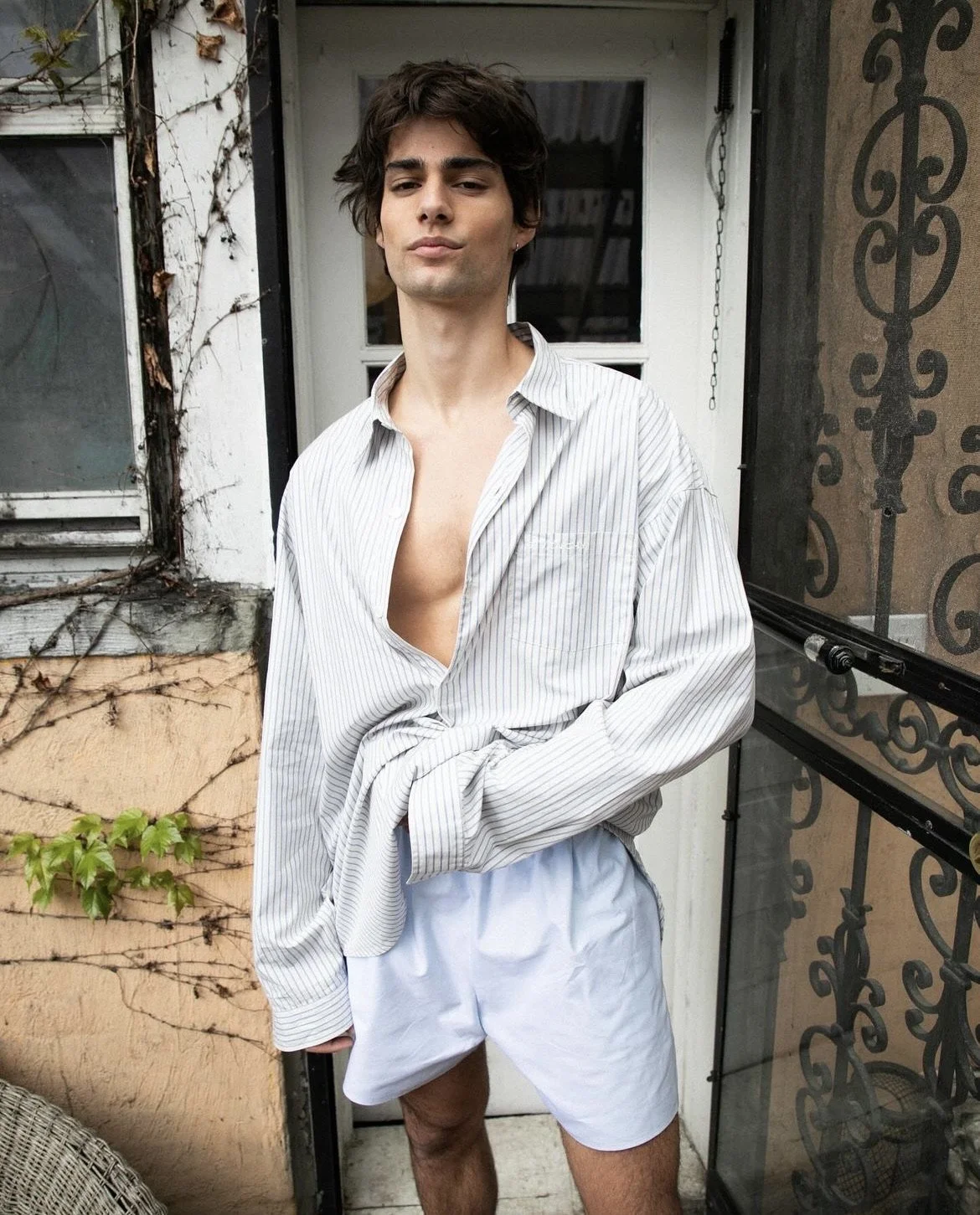A young man stands on a balcony wearing a partially unbuttoned striped shirt and white shorts, with a background of a weathered wall, a window, and decorative black ironwork.