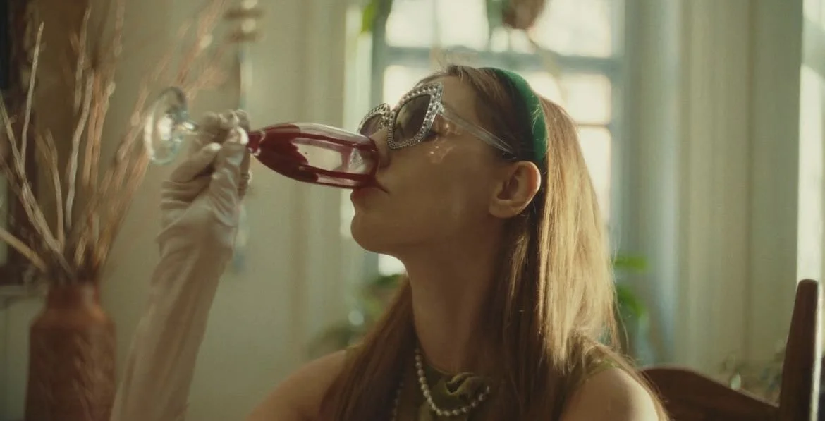 Woman wearing sunglasses and a headband drinking from a large wine glass in a bright room with green plants.
