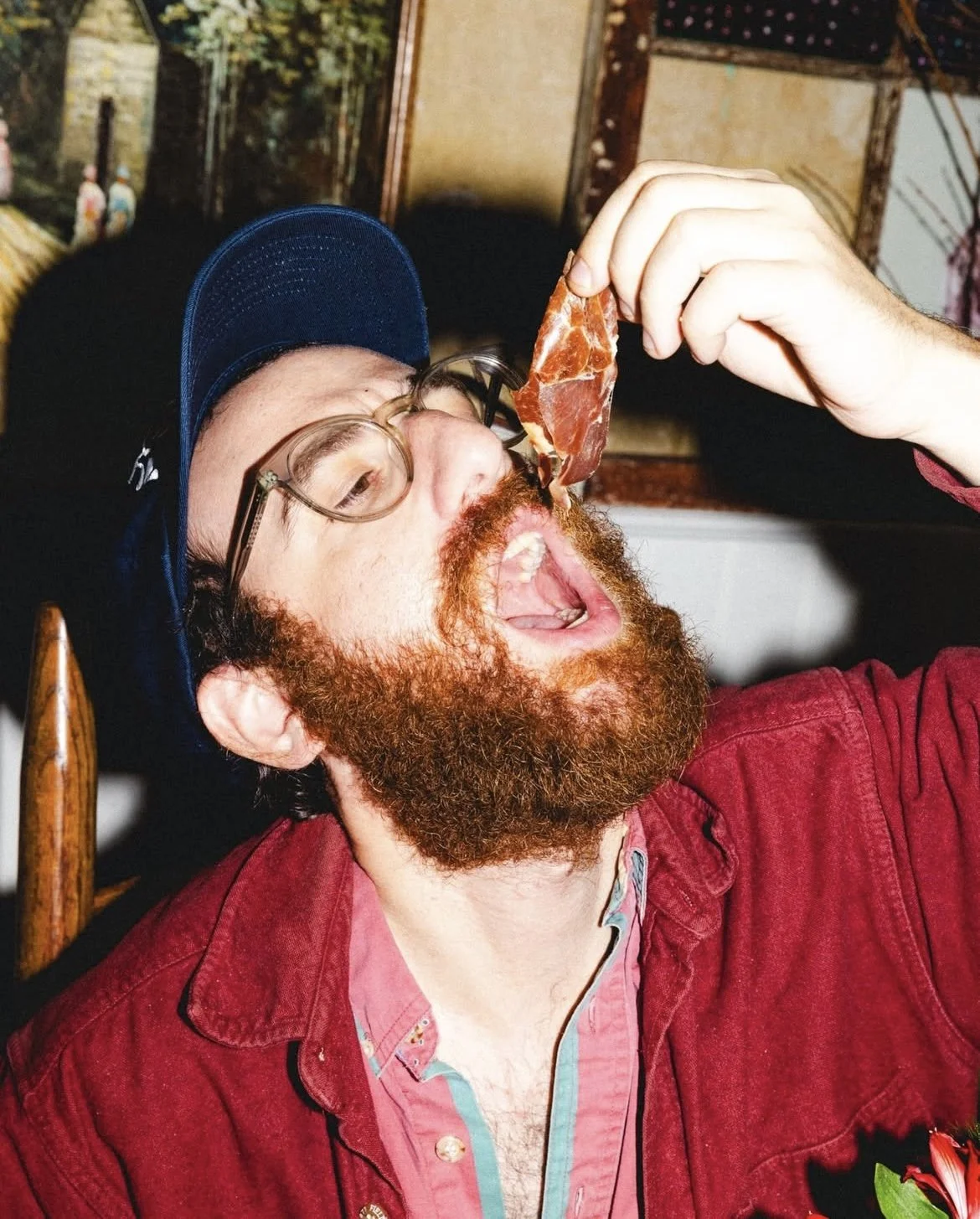 Man with red hair and beard wearing glasses and a blue cap, sitting in a restaurant, opening his mouth as someone holds a slice of cured meat near his open mouth.