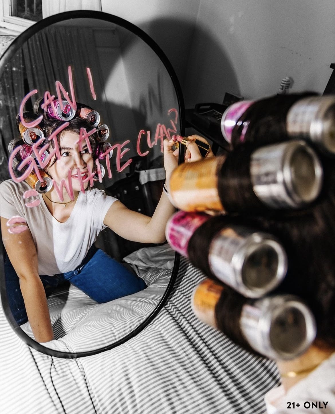 Young woman with curlers in her hair, writing with a marker on a round mirror that says ''Can I get a wet wipe?'', as seen through a reflection from a rack of spray bottles in the foreground.