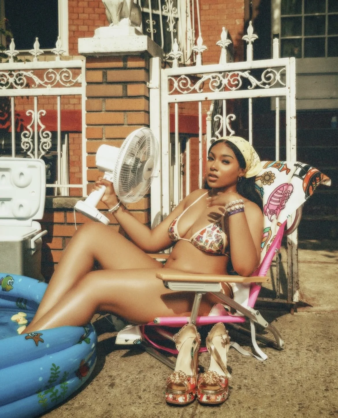 A woman in a bikini sitting on a lawn chair outside, holding a fan, with a pair of sandals on the ground in front of her, and a small inflatable pool nearby. She is surrounded by a brick wall and a white metal fence.