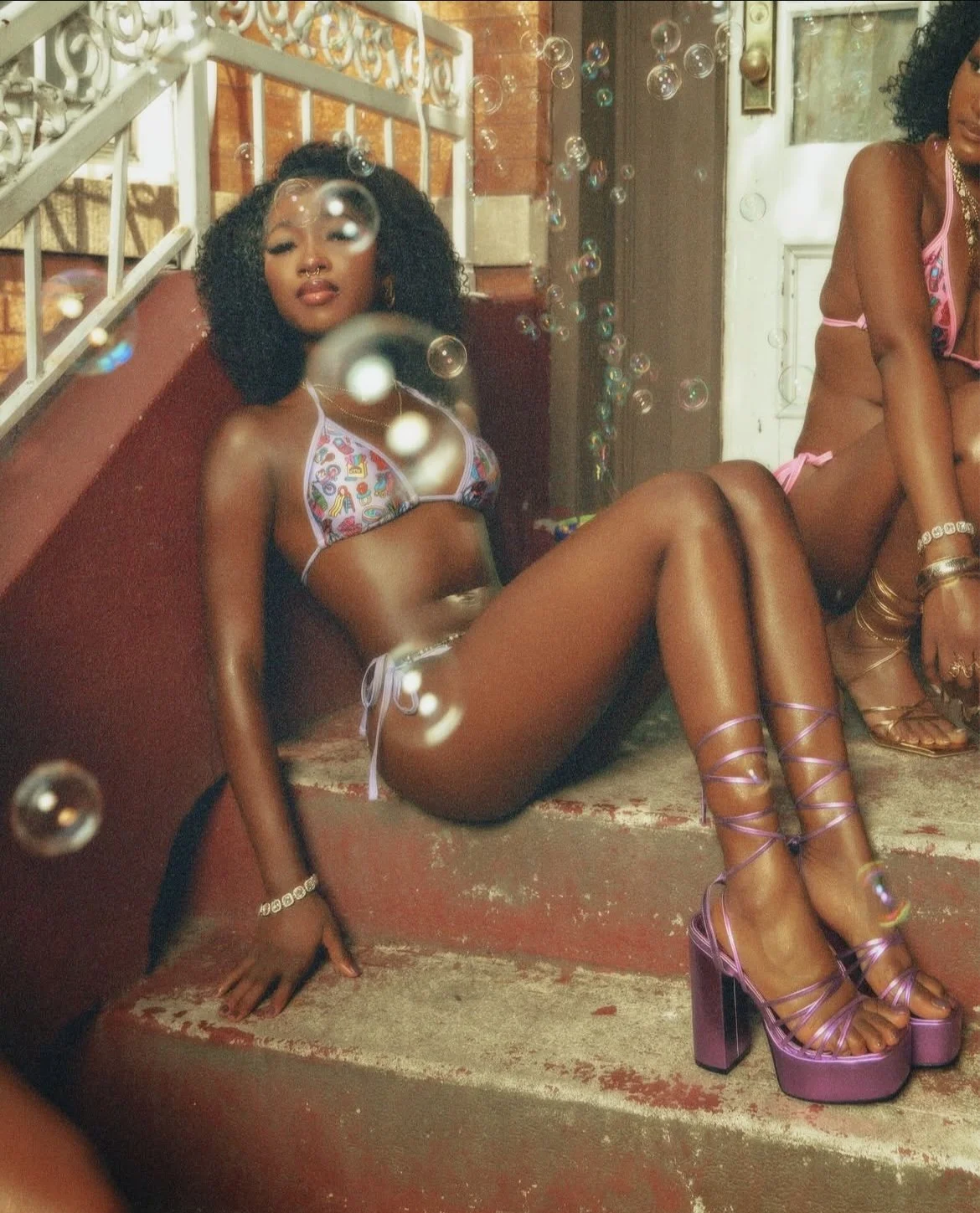 A woman with curly black hair, wearing colorful lingerie and purple high heels, sitting on stairs surrounded by floating soap bubbles.