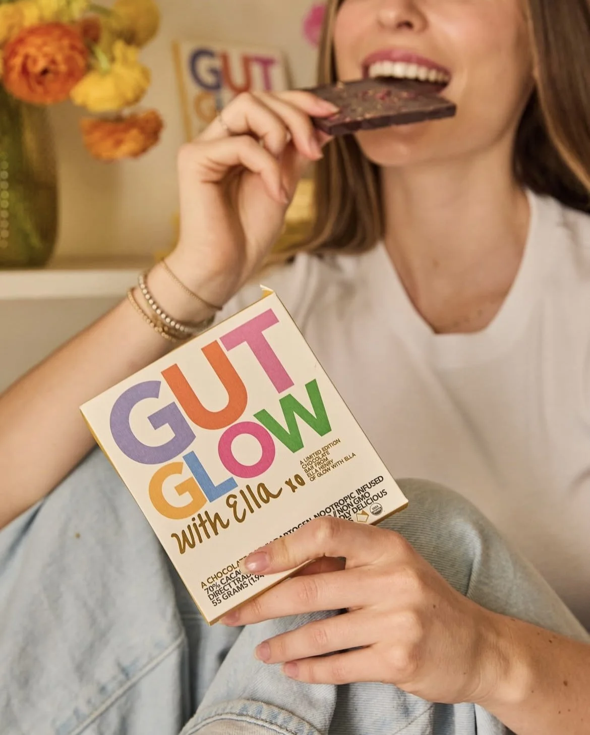 A woman is sitting and smiling while eating a chocolate bar and holding a box of 'GUT GLOW' with colorful text, with a vase of flowers and a decorative sign in the background.