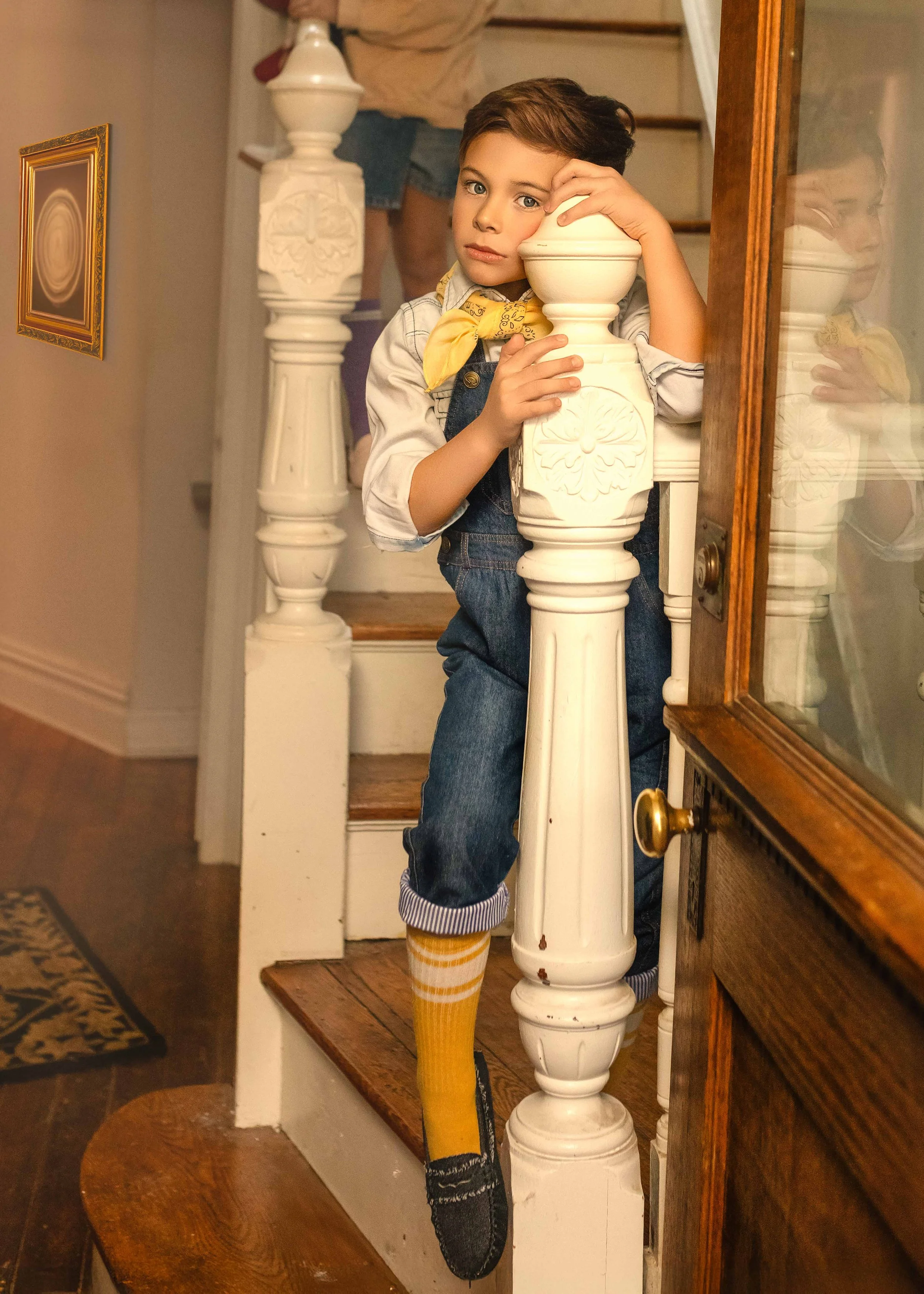 A young boy with brown hair and blue eyes leaning on a staircase railing in a vintage-style house. He's wearing blue jeans, a yellow bandana, and a white shirt, with his foot in a sock and slipper. The staircase has white balusters and wooden steps, 