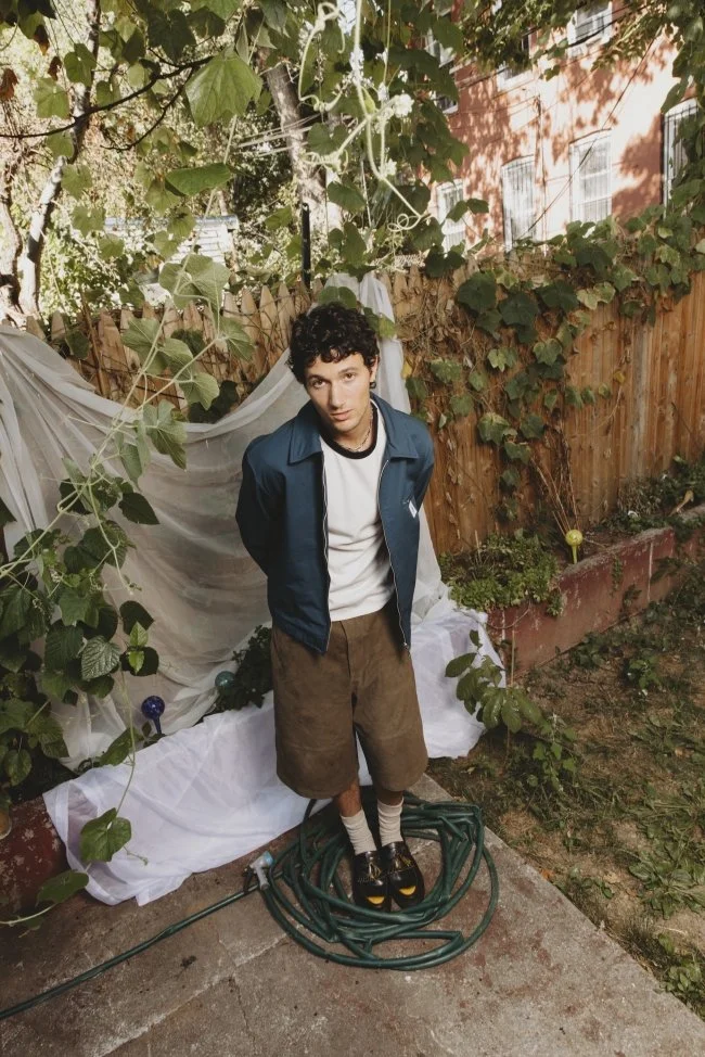 A young man with curly dark hair standing in a backyard with a wooden fence, green plants, and white fabric draped over the fence. He is wearing a blue jacket, white T-shirt, brown shorts, striped socks, and platform shoes, with garden hoses coiled a