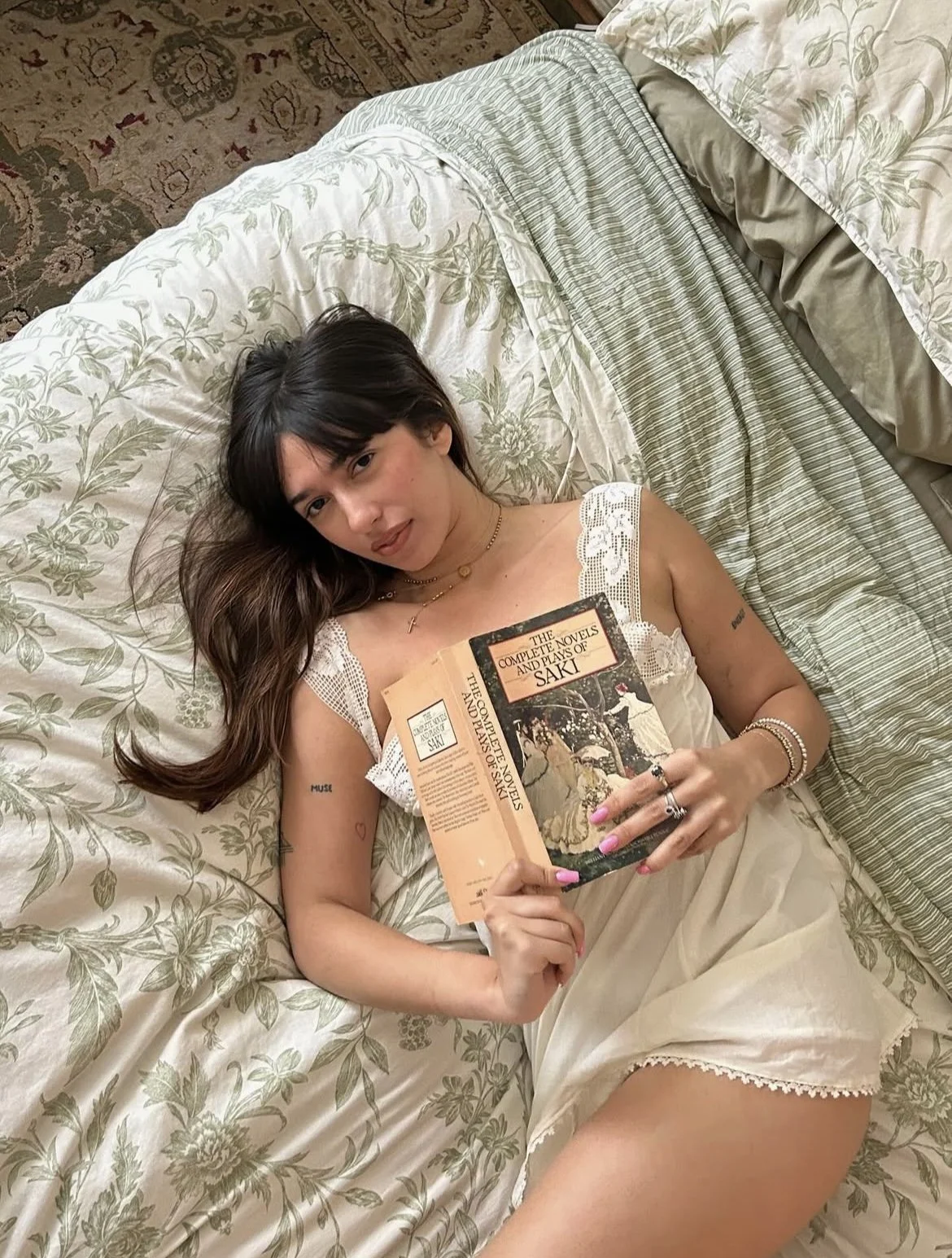 A young woman with dark hair lying on a bed, holding a book titled 'The Complete Novels and Plays of Saki.' She is wearing a white lace detail dress, jewelry including rings, bracelets, and necklaces, and has small tattoos on her arm.