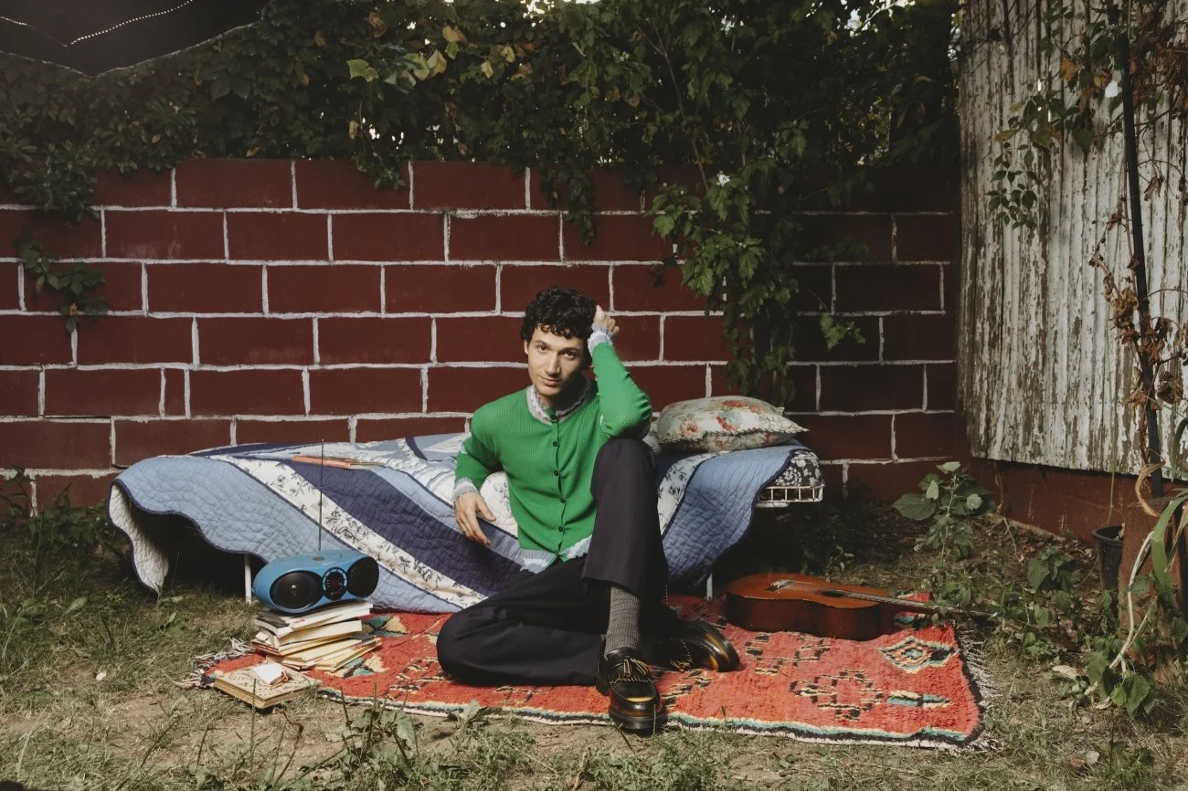 A person sitting on a small bed outdoors with a brick wall and wooden fence in the background. The person is wearing a green sweater, black pants, and black shoes, with a guitar resting beside them, and a portable speaker and books on the ground near