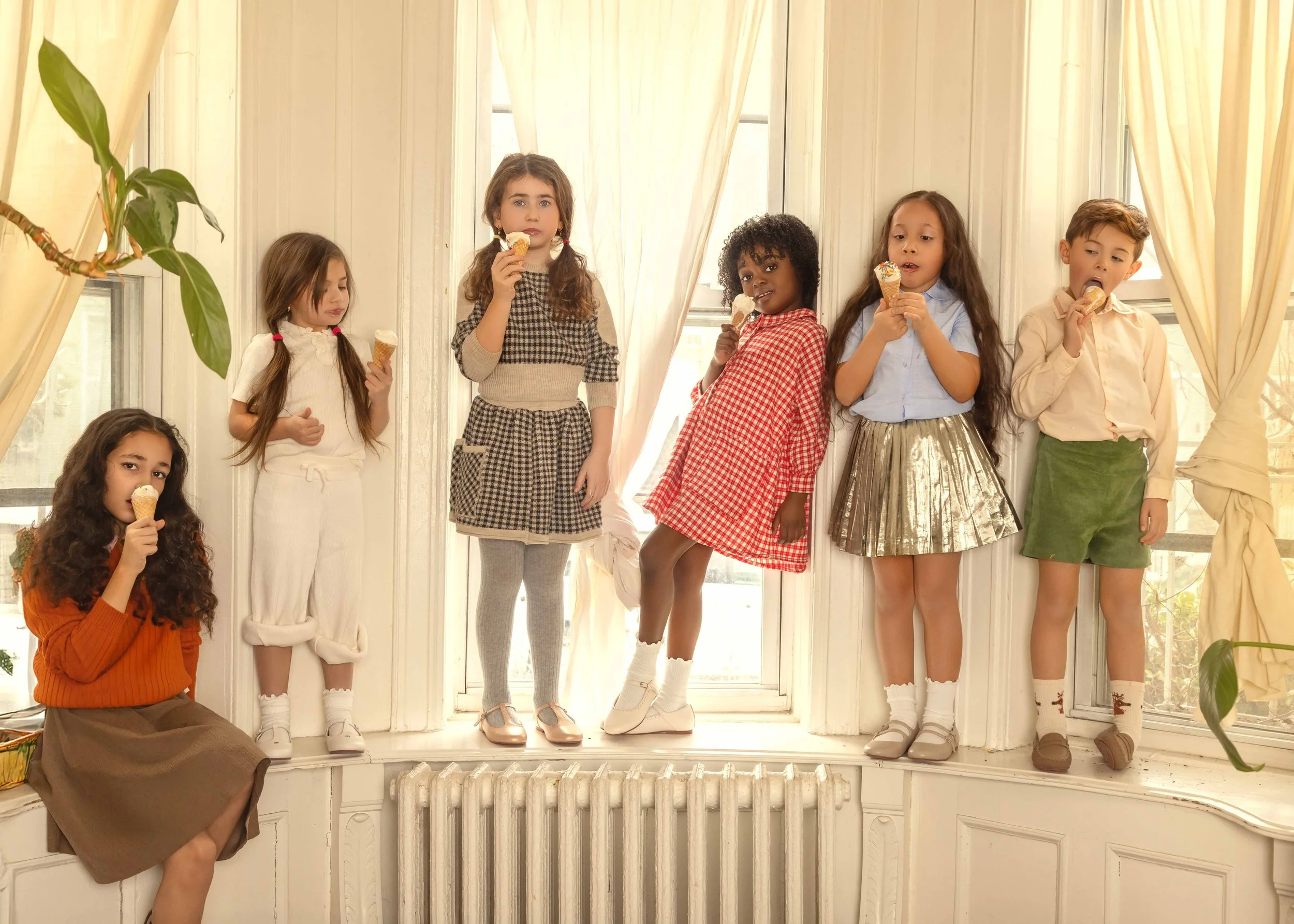 Seven children with diverse ethnicities and dressed in colorful clothing, eating ice cream cones while standing on a large window seat in a bright room.