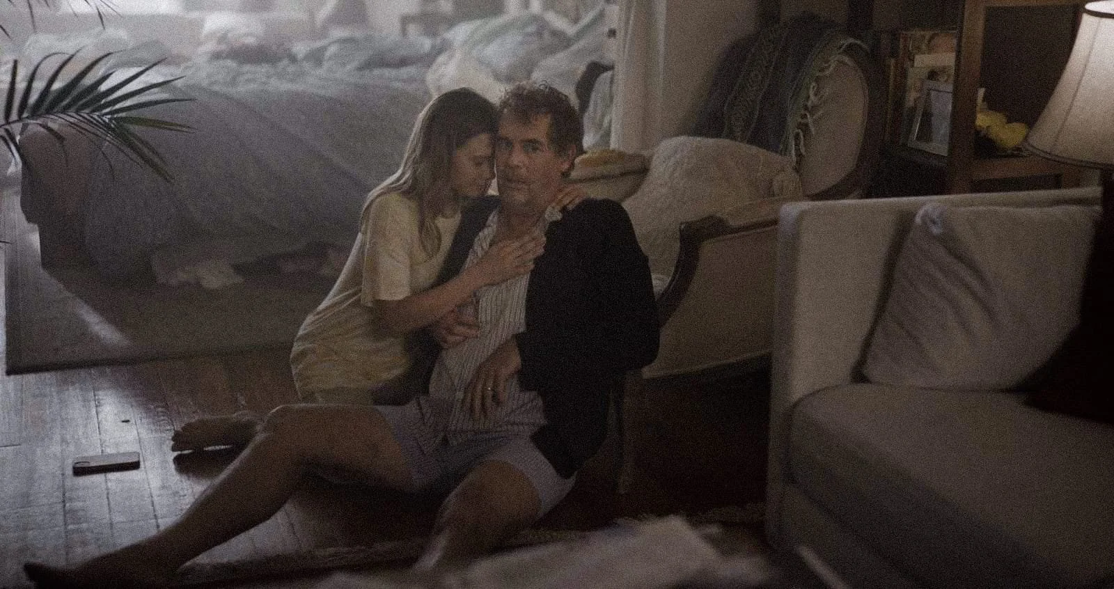 A man sitting on the floor indoors, holding a young girl on his lap, with another woman leaning in close. The setting shows a cozy room with sofas, a table lamp, and a large picture or tapestry of a bed and flowers in the background.