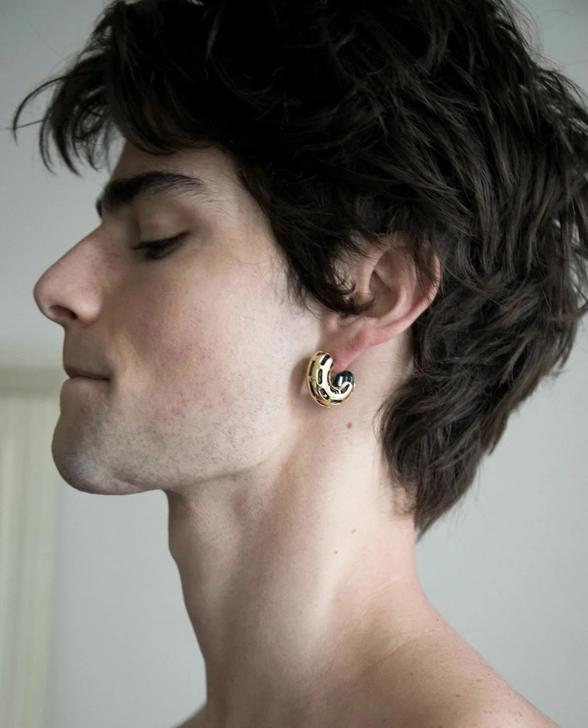 Close-up of a young person with short, wavy dark hair wearing a gold and black ringed earring on the left ear, looking to the left with eyes closed.
