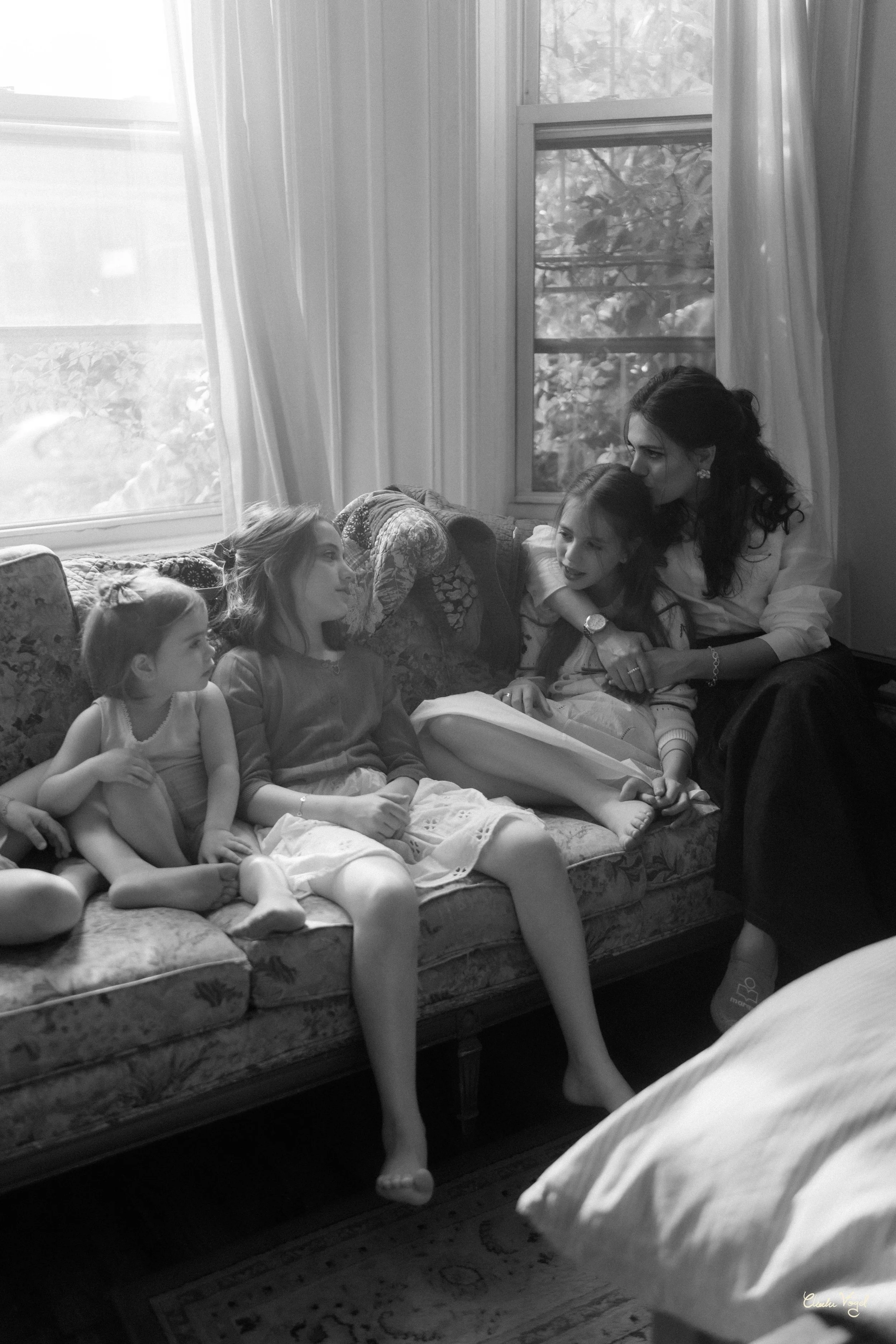 A woman and three young girls sitting on a floral-patterned couch in a room with large windows and curtains, engaged in conversation.