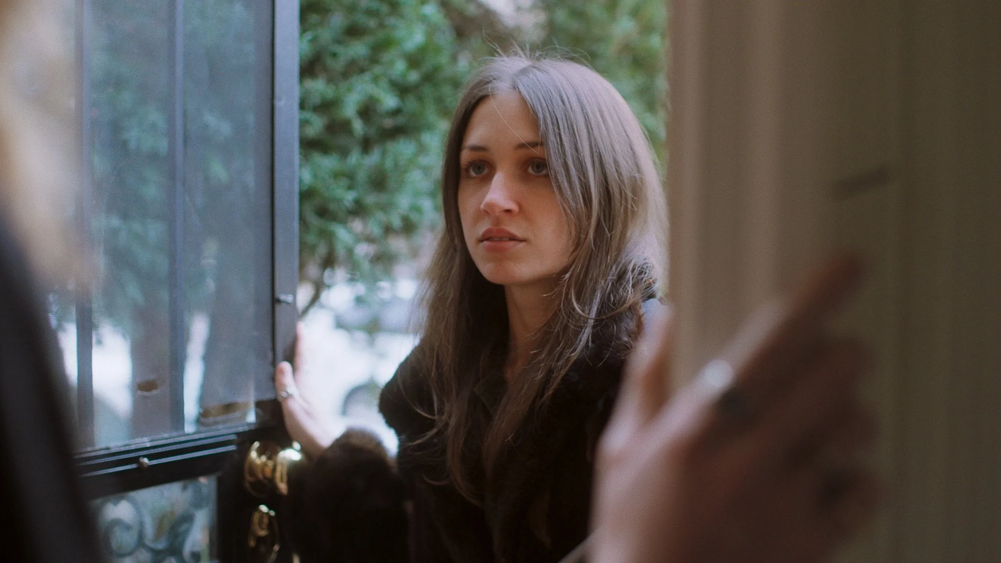 A woman with long, wavy hair standing at a door, looking serious or concerned, with her hand on the door frame.