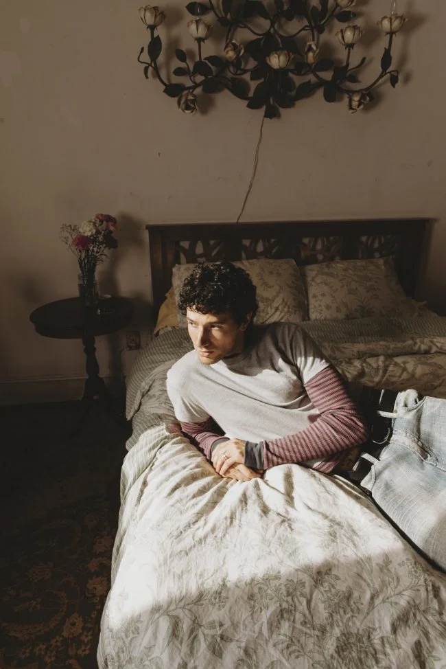A young man lying on a bed in a dimly lit room, with a floral bedspread, a wooden headboard, a nearby round table with a vase of flowers, and an ornate ceiling light fixture.