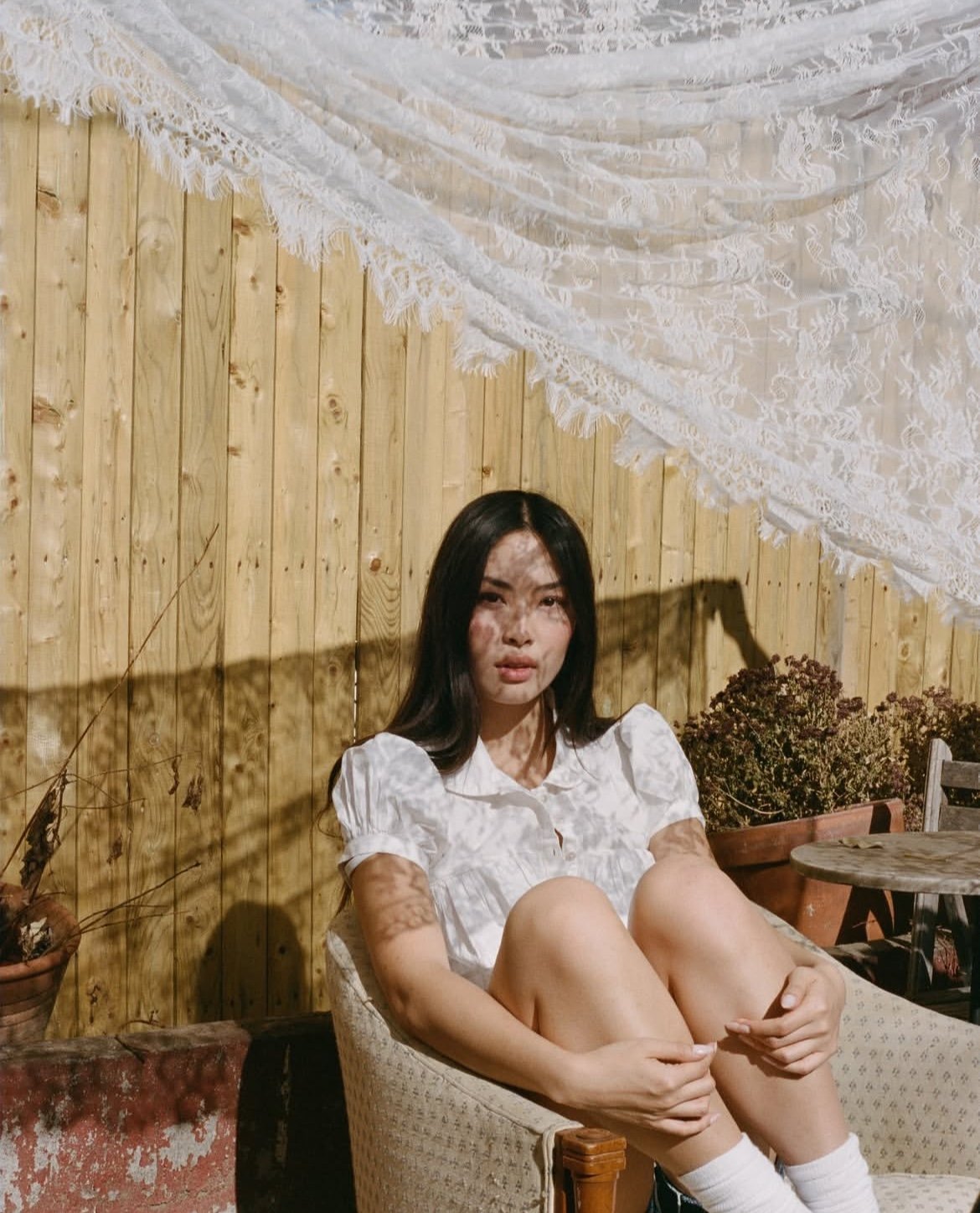 A woman with dark hair, wearing a white blouse, sitting on a cushioned chair outdoors against a wooden wall. Sunlight casts shadow patterns on her face and arms.