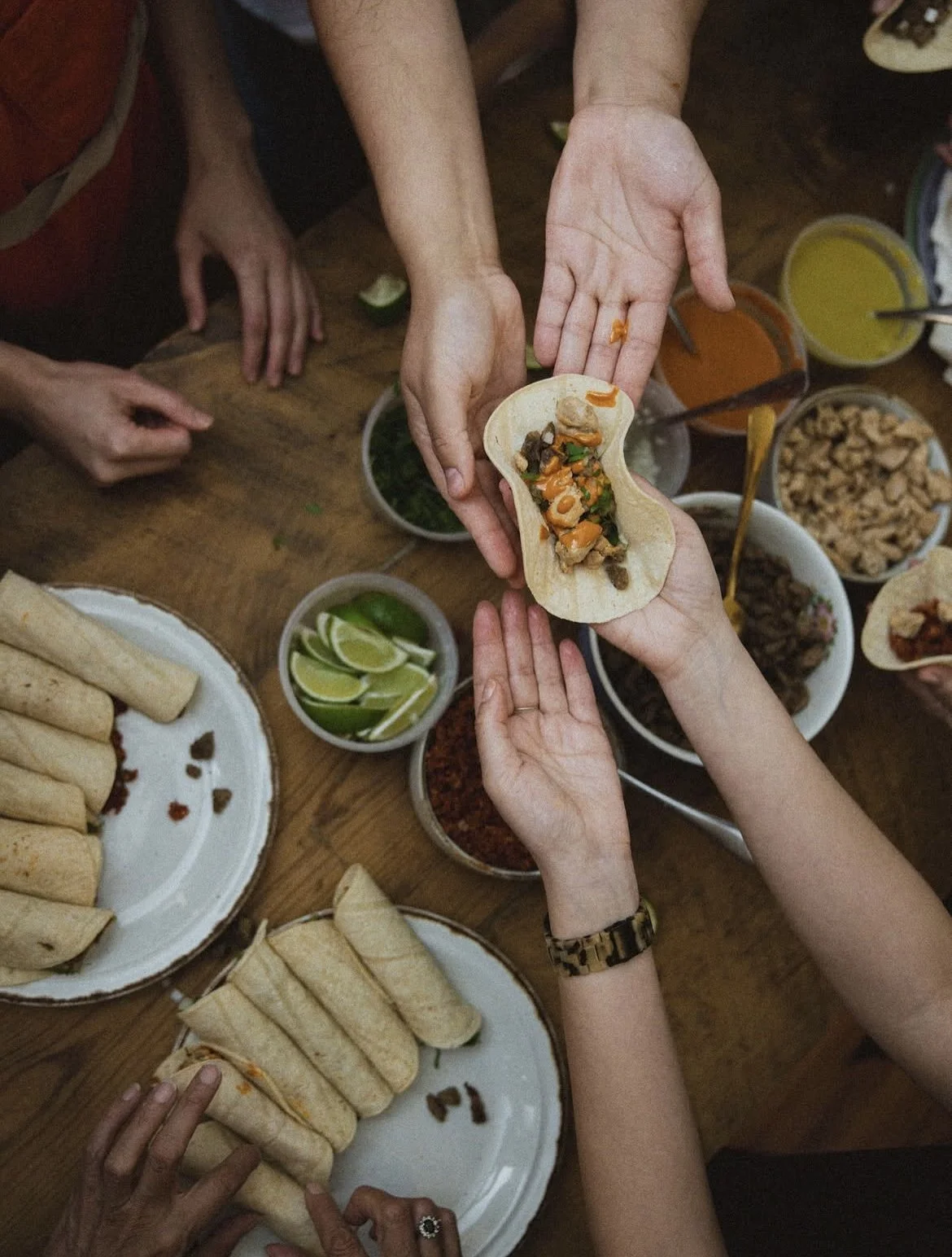 People sharing street tacos surrounded by plates of rolled tortillas, lime wedges, and various sauces.