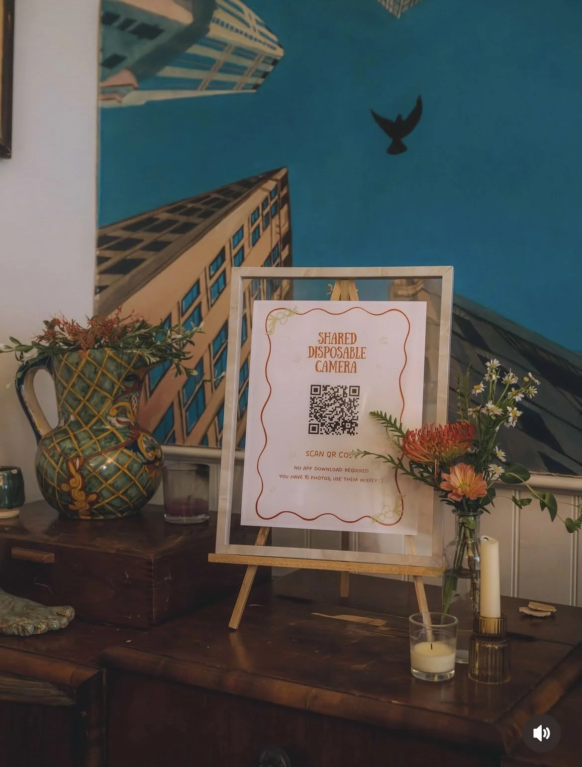 Sign for shared disposable camera with QR code, titled 'Shared Disposable Camera'; decorative vase with flowers, candle, and plants on a wooden surface, with a mural of tall buildings and sky with a bird in the background.